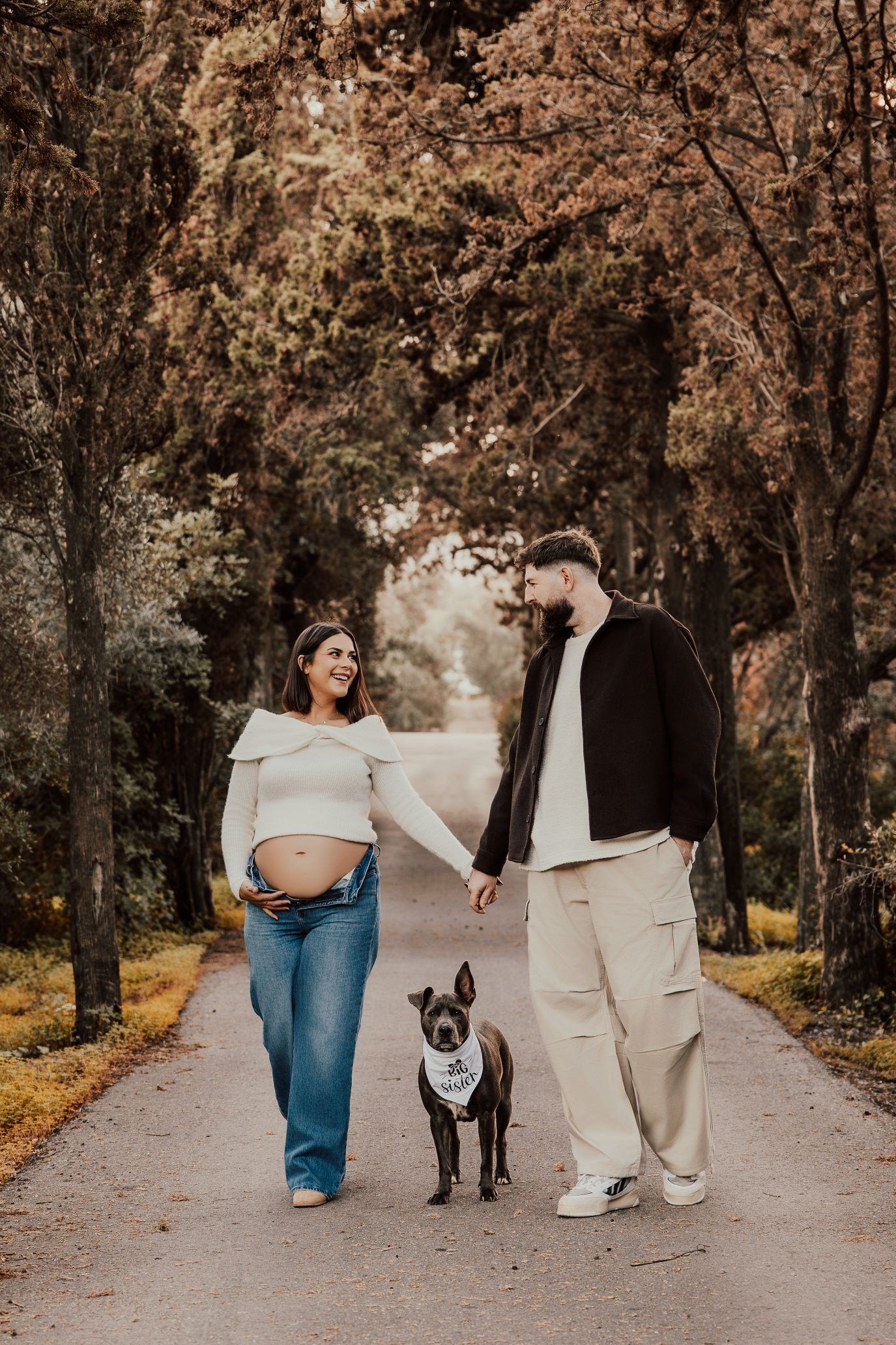 Una pareja pasea a su perro por un sendero arbolado, tomados de la mano bajo una suave luz otoñal.