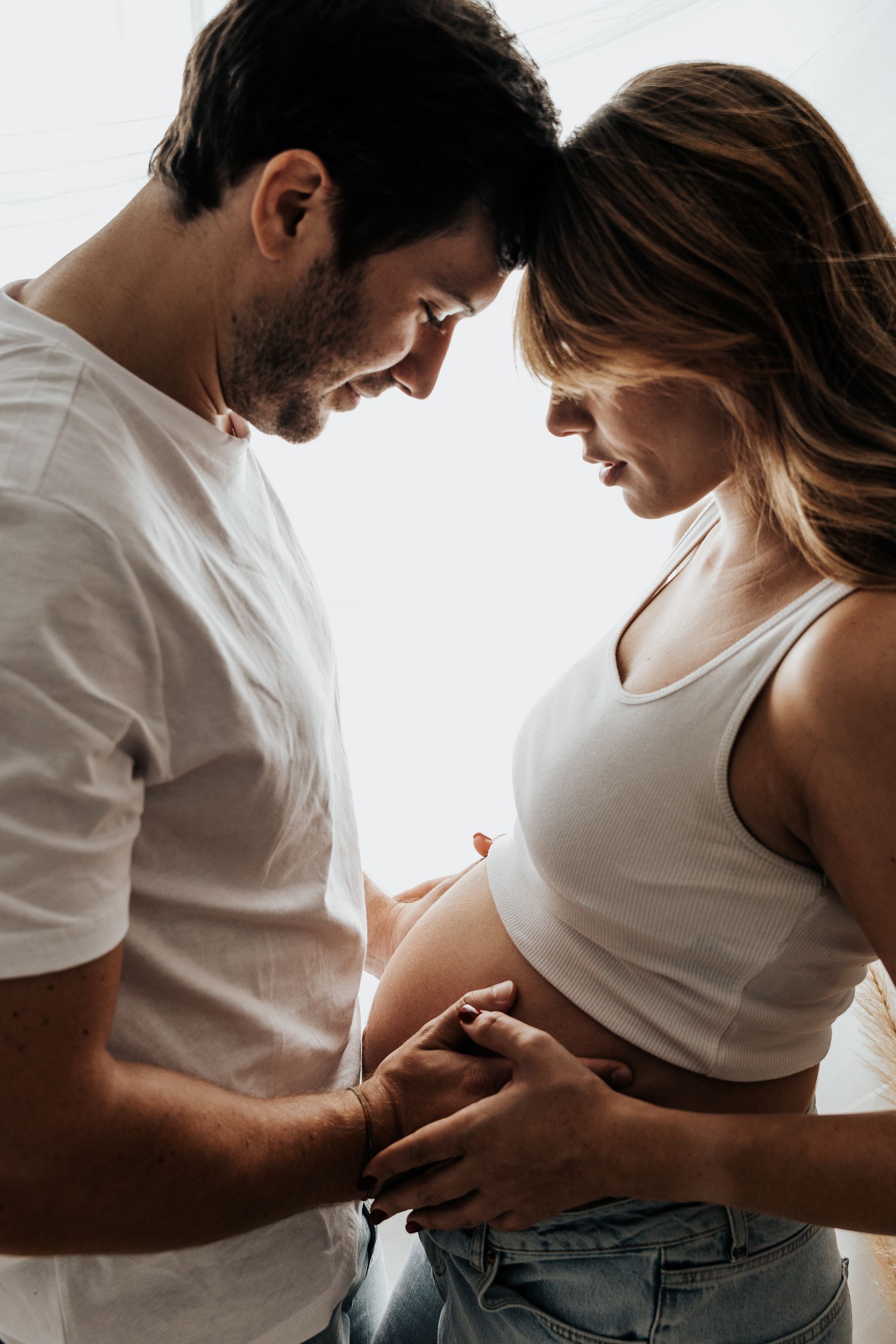 Una pareja se toma de las manos con delicadeza y se toca la frente, ambos con camisas blancas sobre un fondo brillante.