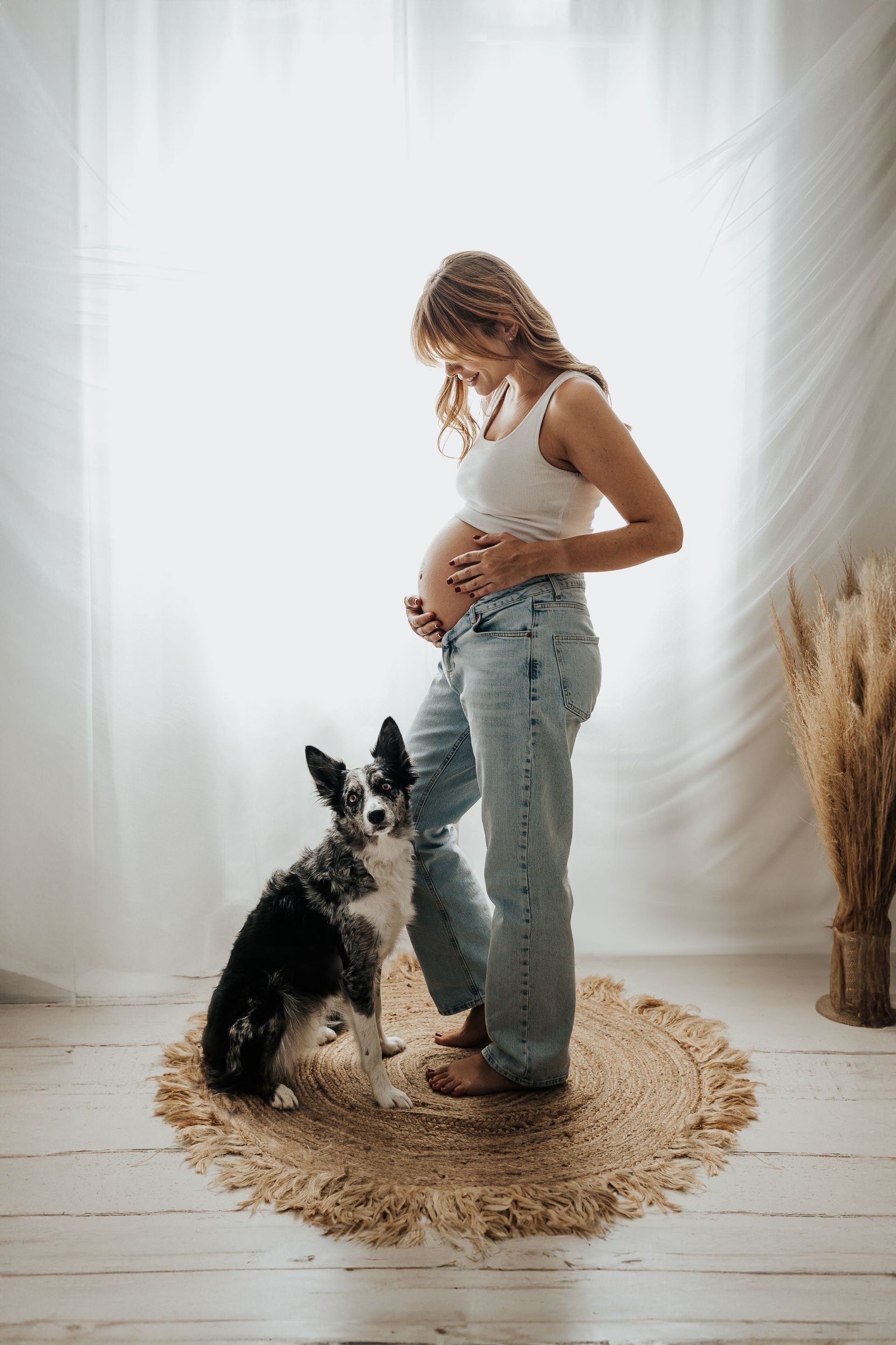 Mujer embarazada con blusa blanca y jeans de pie junto a un perro blanco y negro sobre una alfombra redonda junto a una ventana luminosa.