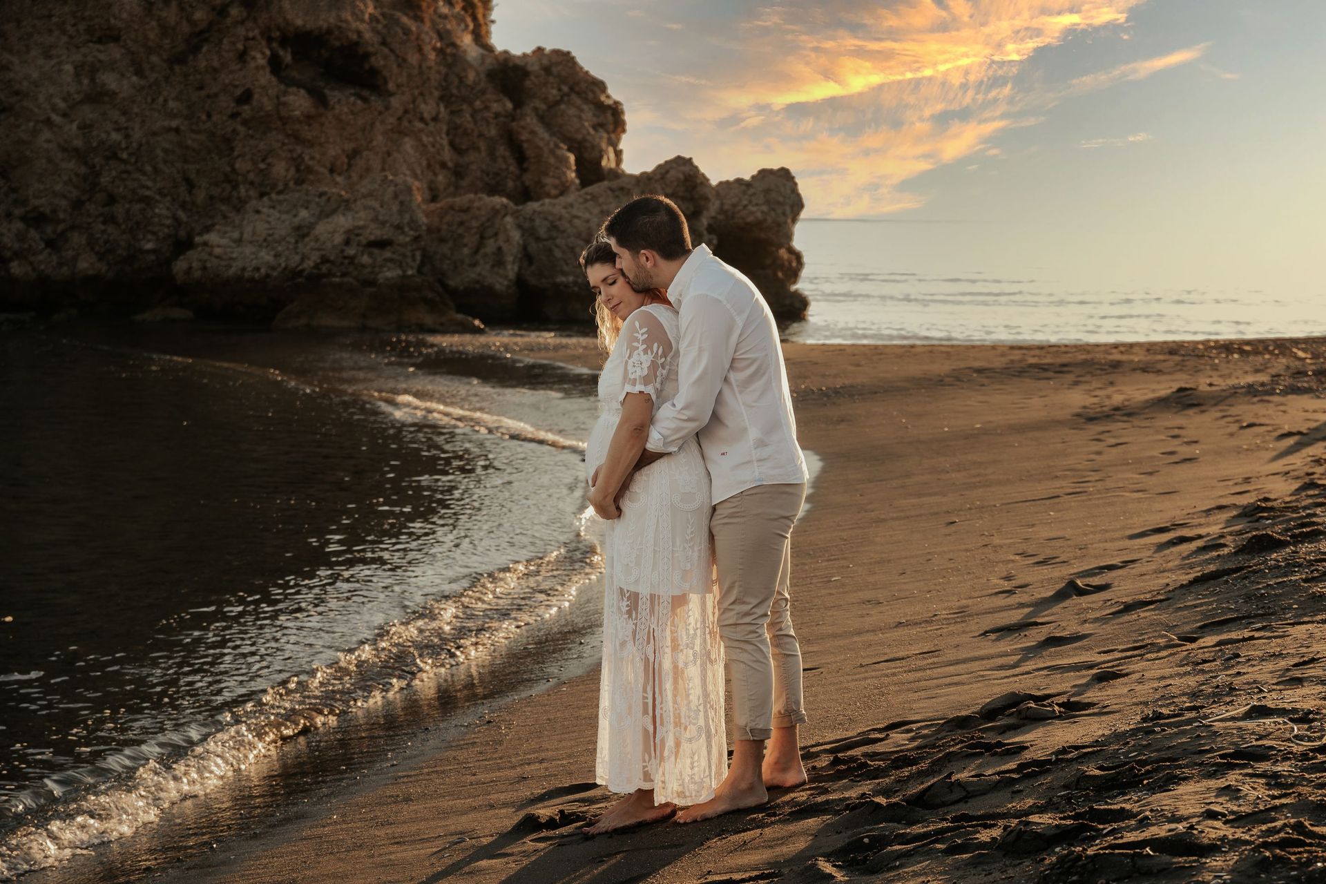 Una pareja vestida de blanco se abraza en una playa al atardecer, junto a acantilados rocosos y olas suaves.