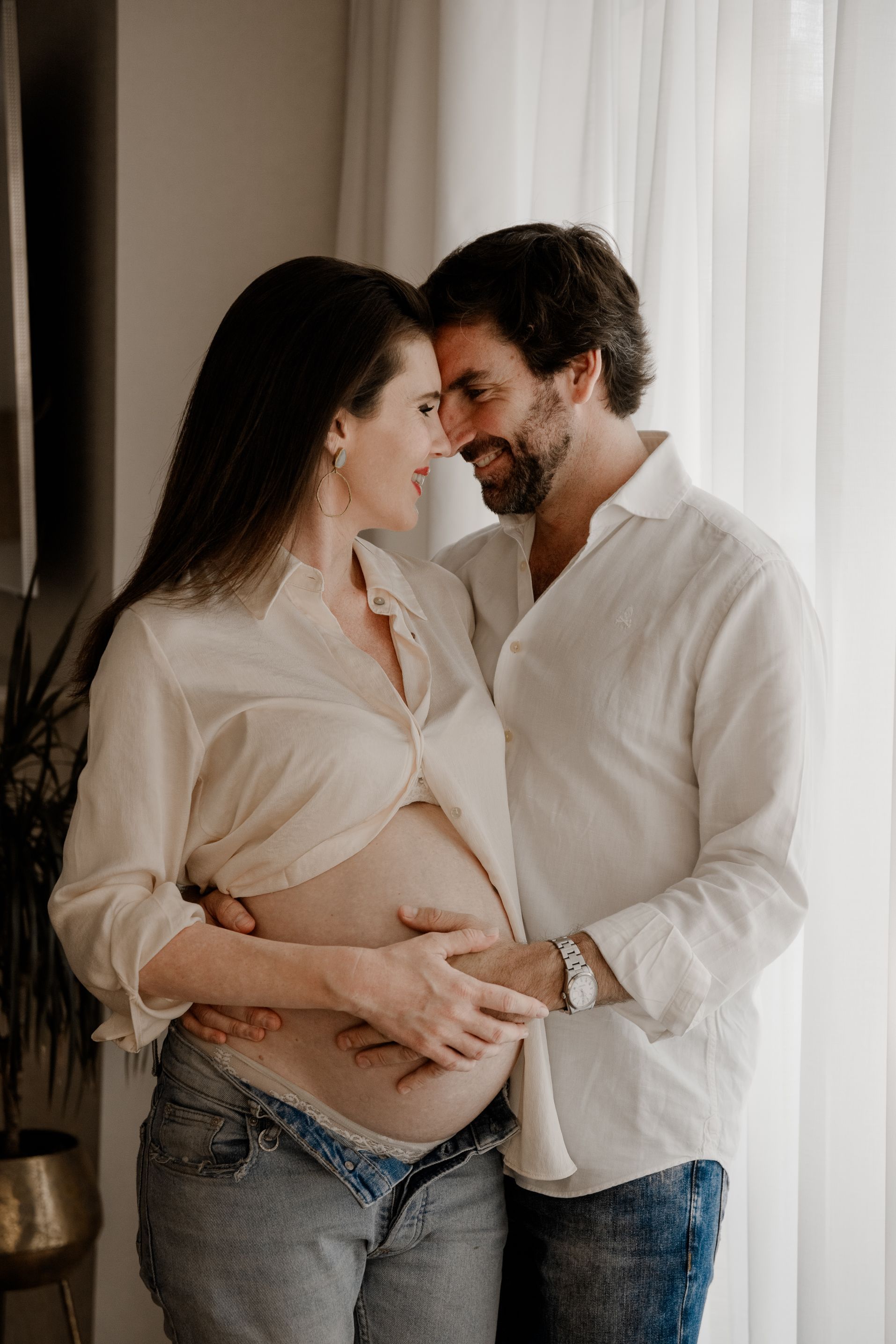 Una pareja sostiene con delicadeza la barriga de una mujer embarazada junto a una ventana, sonriendo bajo una suave luz natural.