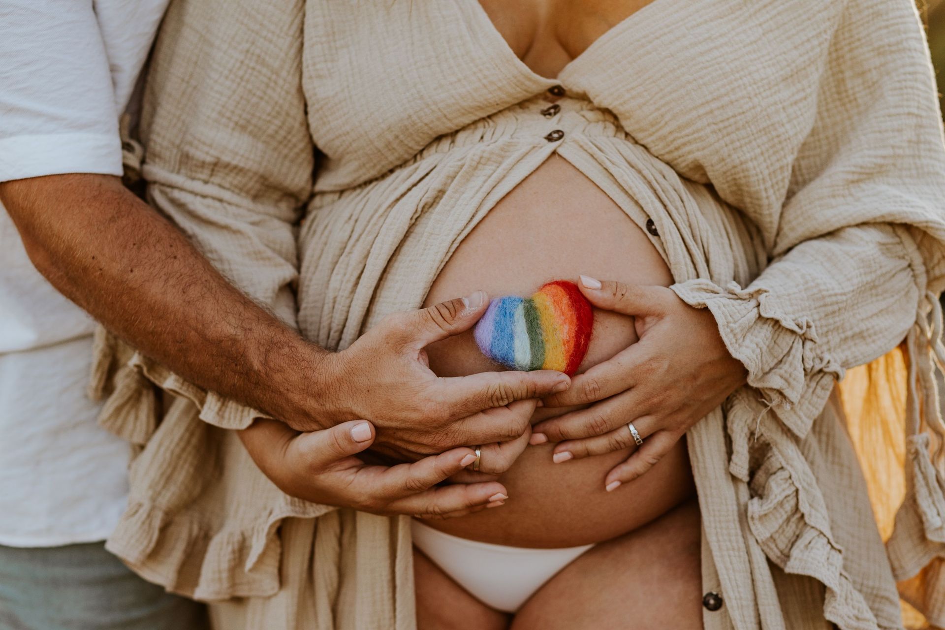 Manos sosteniendo una barriga de embarazada pintada con un arcoíris en un vestido beige.