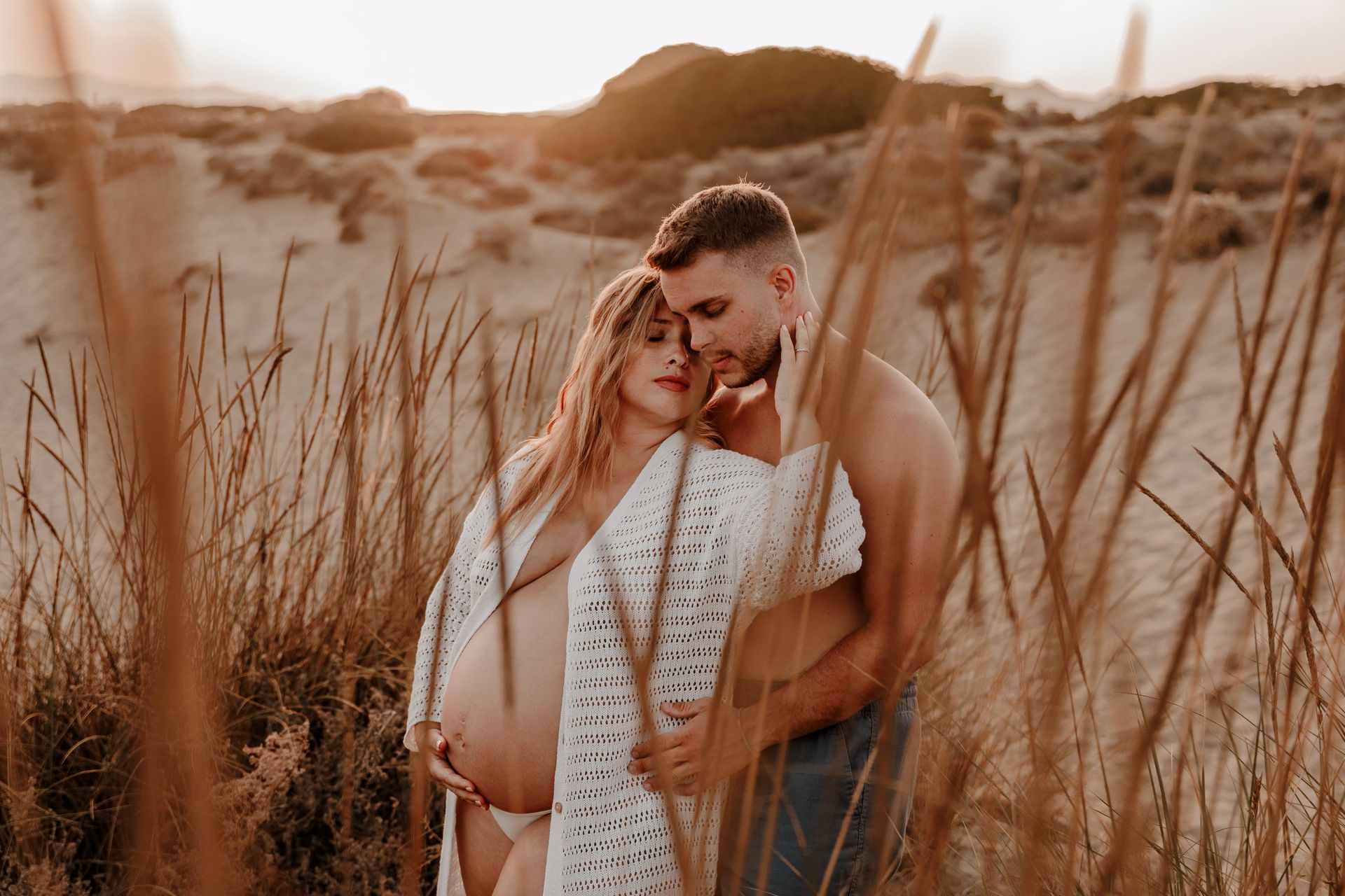 Pareja abrazándose entre la hierba alta de la playa al atardecer, con una cálida luz dorada.