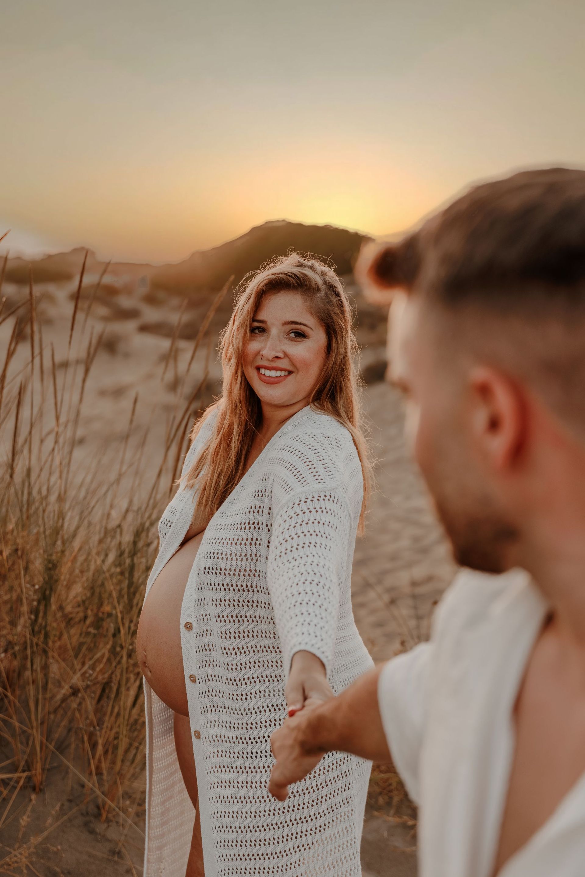 Mujer embarazada con un vestido blanco sonriendo al atardecer, tomada de la mano de su pareja en un campo de hierba.