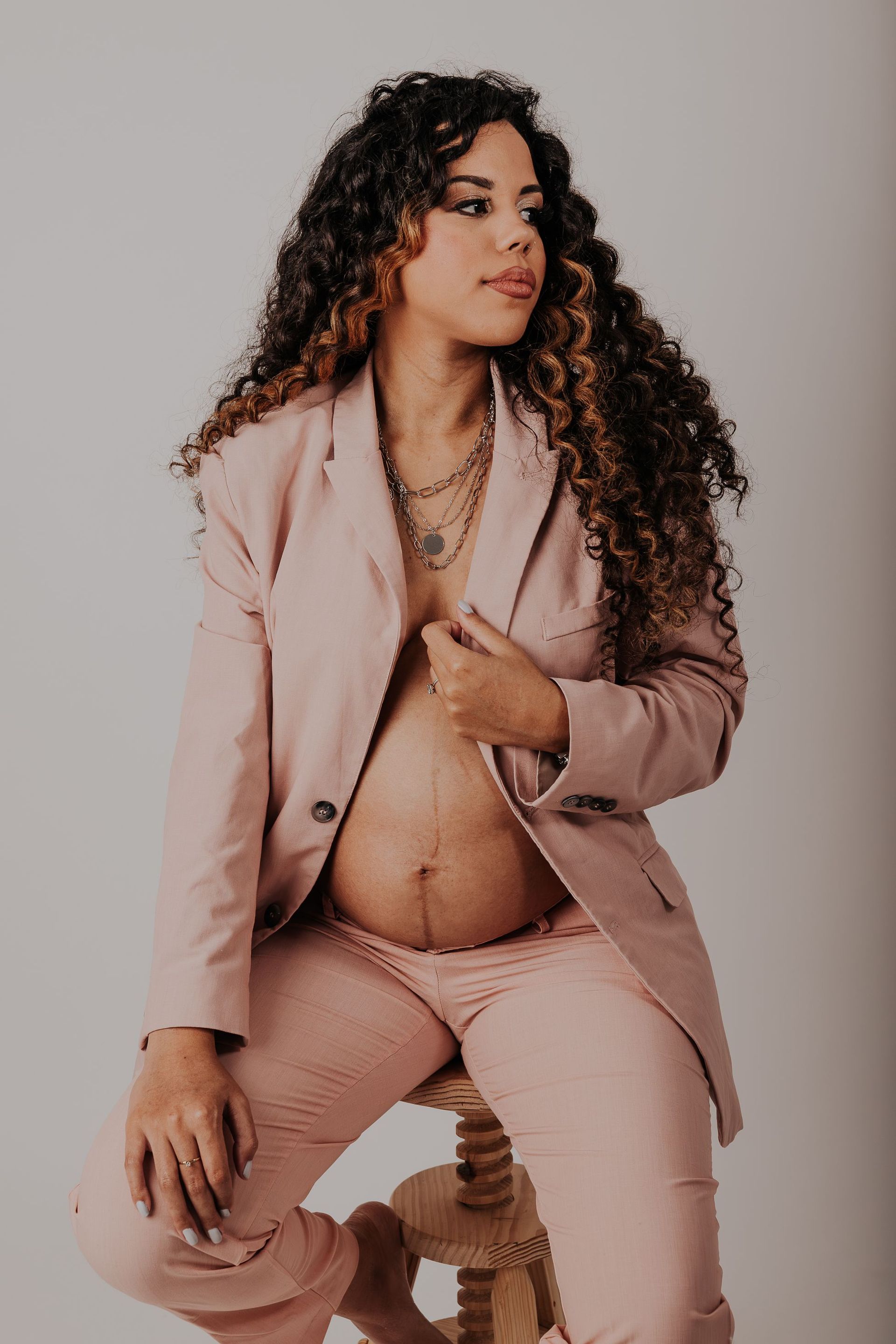 Mujer con traje color rosa empolvado sentada en un taburete, con la chaqueta abierta, sobre un fondo gris liso.
