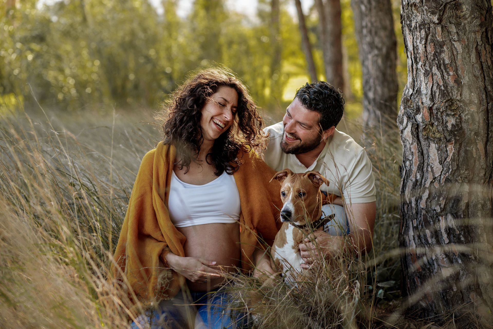 Pareja sonriendo con un perro en la hierba alta entre los árboles, al aire libre en un día soleado.