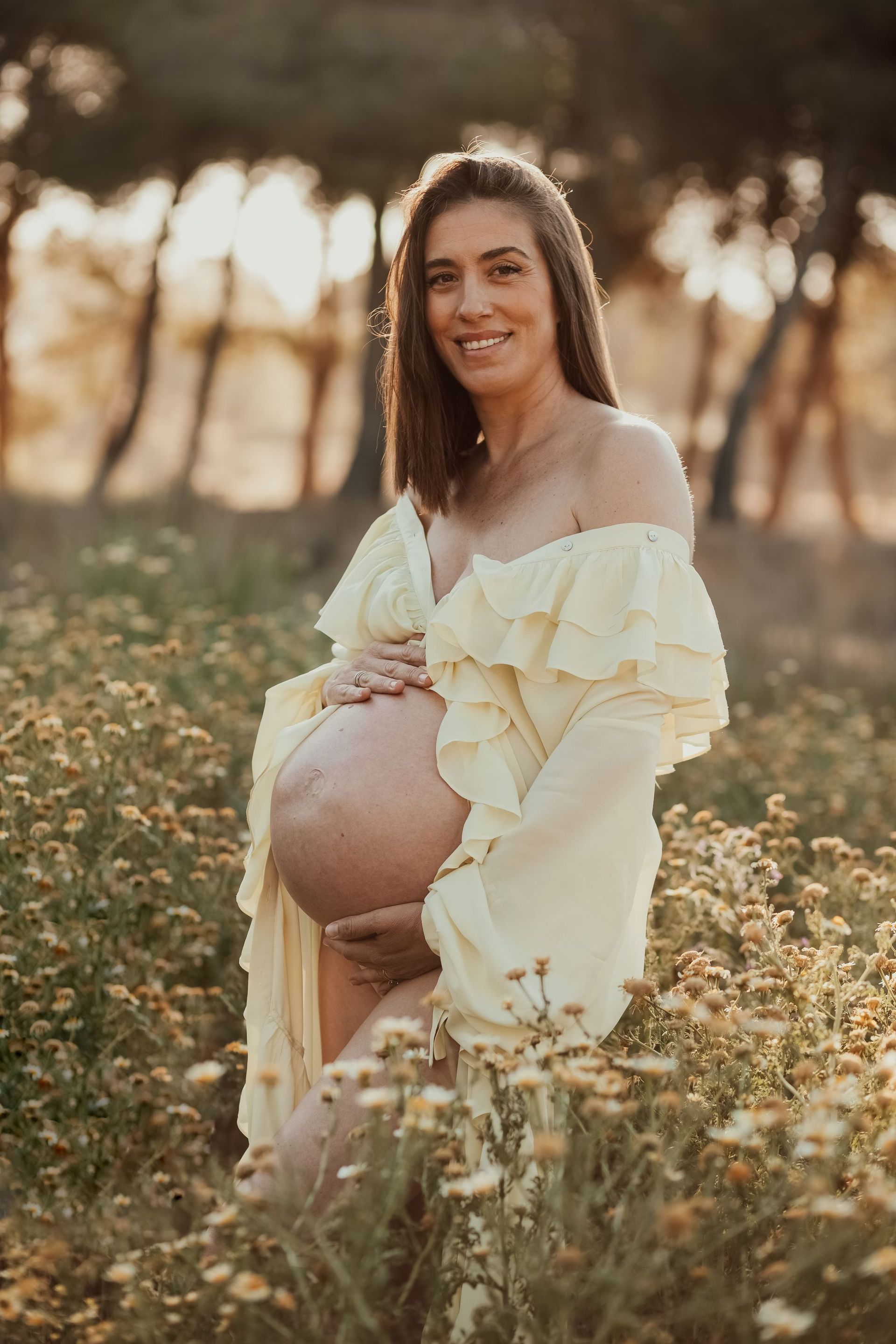 Mujer embarazada con un vestido color crema sentada entre flores silvestres al atardecer, sonriendo a la cámara.