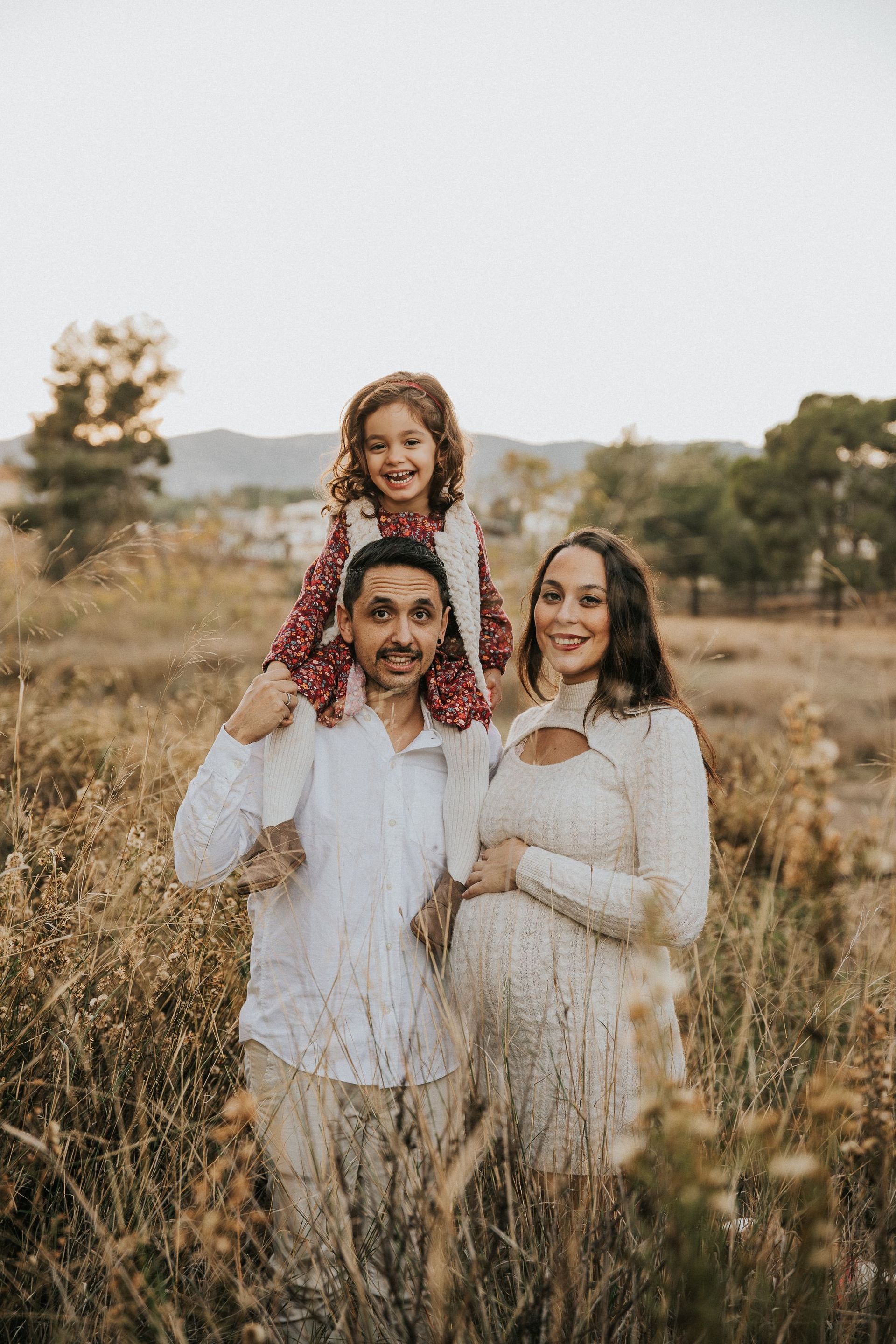 Familia de pie entre la hierba alta, con un niño sobre los hombros de un hombre junto a una mujer sonriente al aire libre al atardecer.
