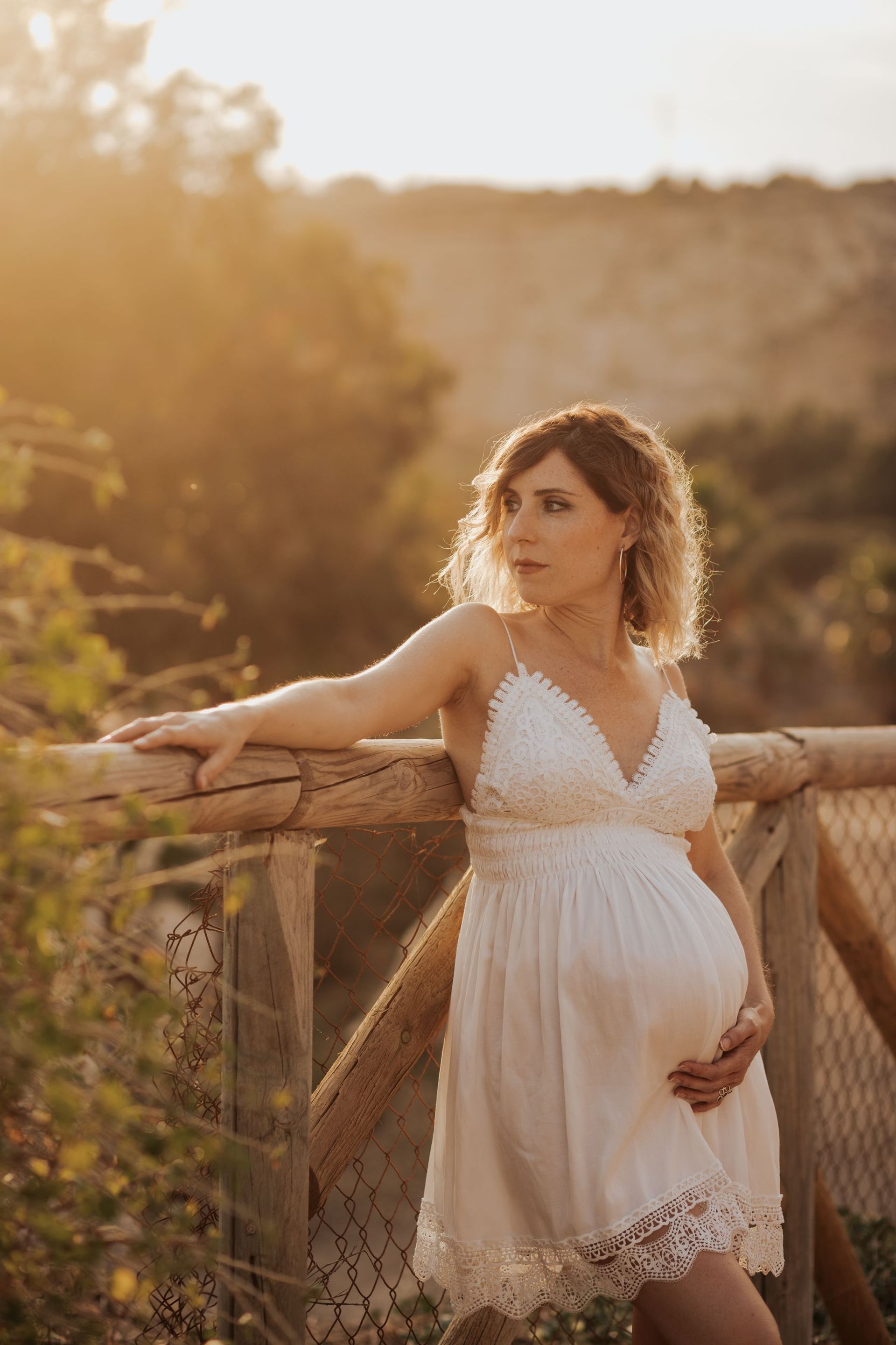 Mujer embarazada con un vestido blanco apoyada en una valla de madera al atardecer, al aire libre en un paisaje rústico.