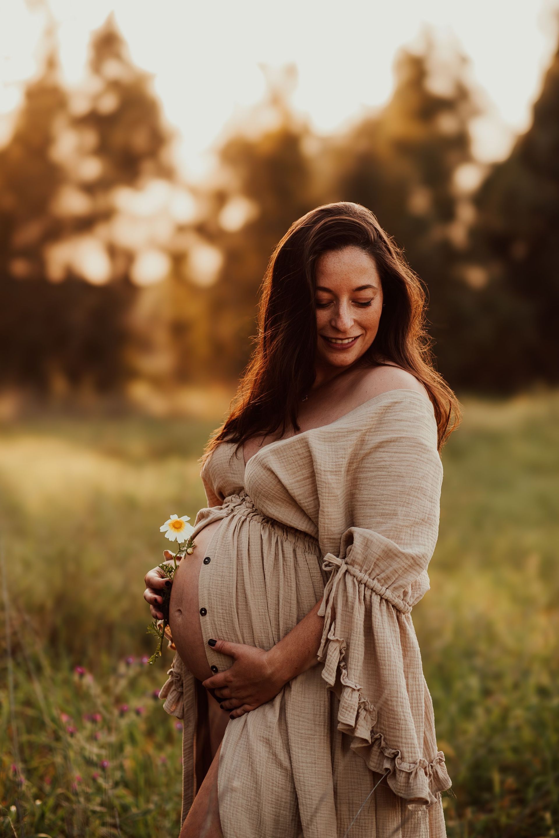 Mujer embarazada con un vestido beige sonriendo en un campo iluminado por el sol al atardecer.
