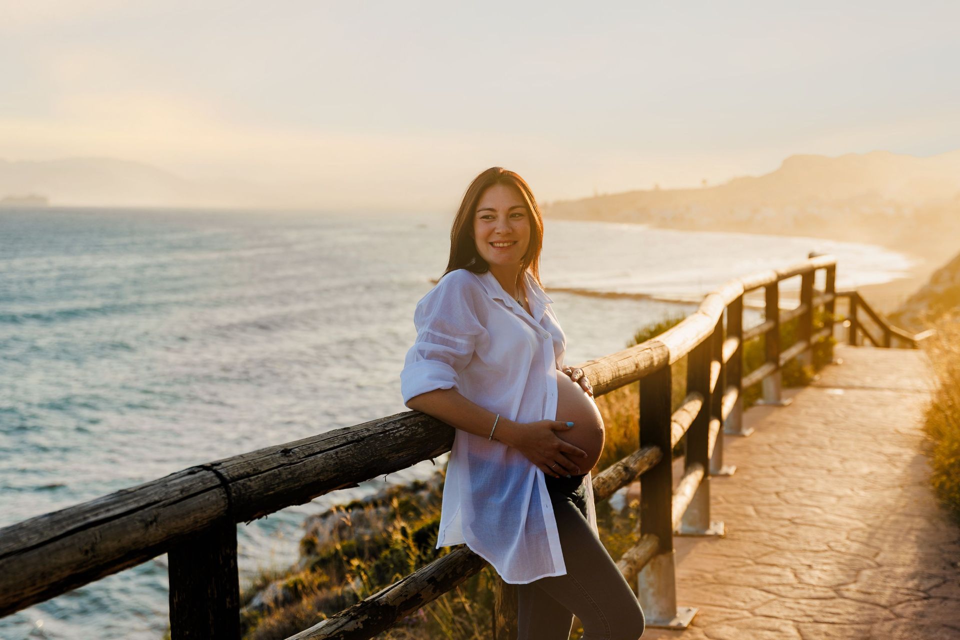 Mujer apoyada en una barandilla junto al mar al atardecer, con el océano y el sendero costero al fondo.