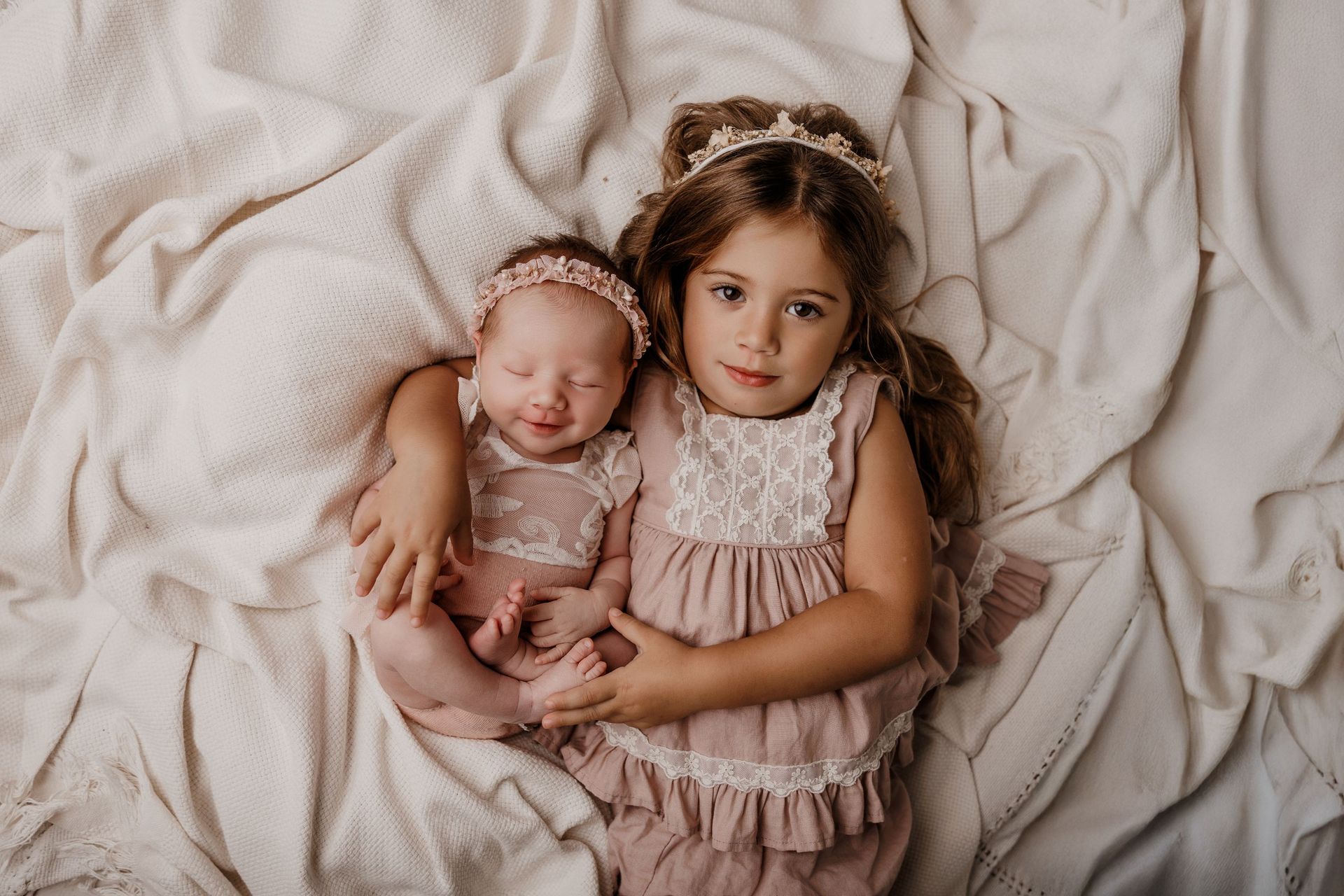 Dos niños pequeños vestidos de rosa yacen acurrucados sobre una manta color crema.