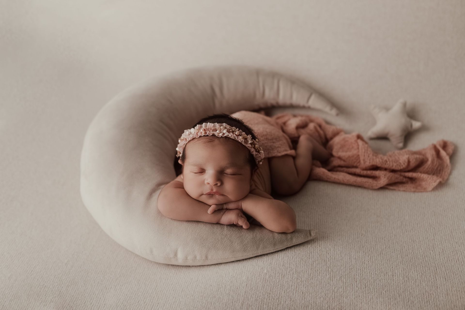 Bebé durmiendo sobre un cojín en forma de media luna color crema, cubierto con una manta rosa en una cama beige.