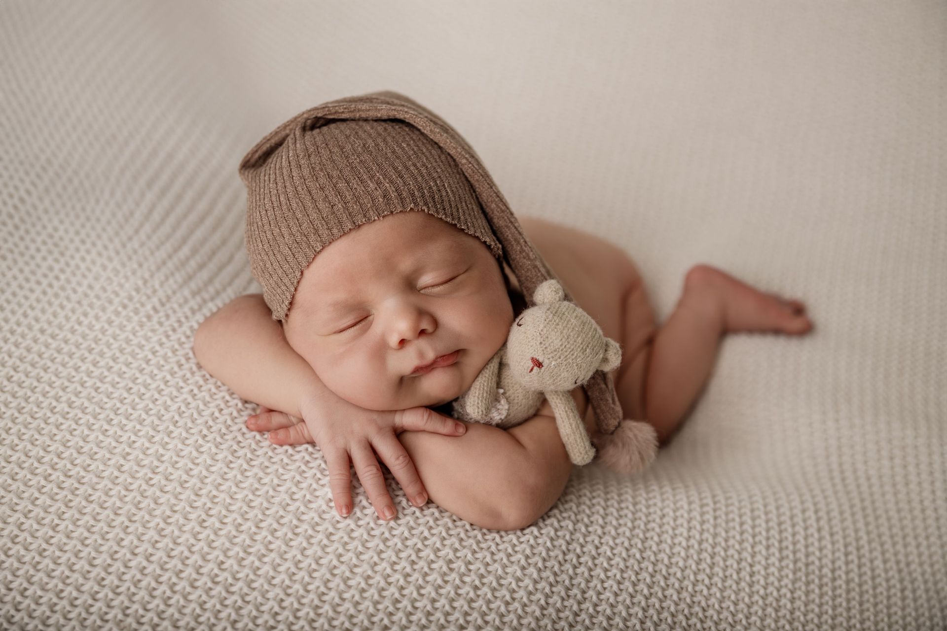 Bebé durmiendo con un gorro de punto marrón, tumbado sobre una suave manta beige con un pequeño osito de peluche.