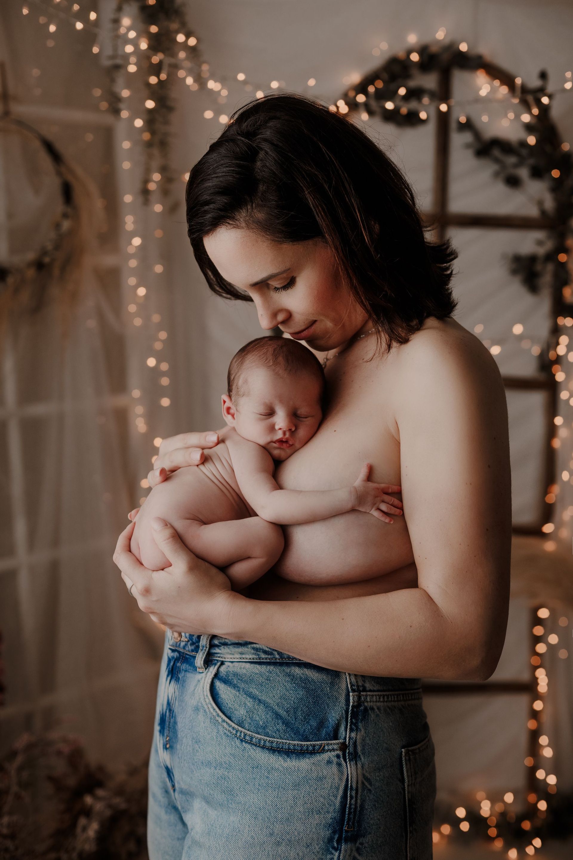 Una mujer sostiene a un bebé dormido en una habitación tenuemente iluminada con guirnaldas de luces y una caja de regalo envuelta cerca.