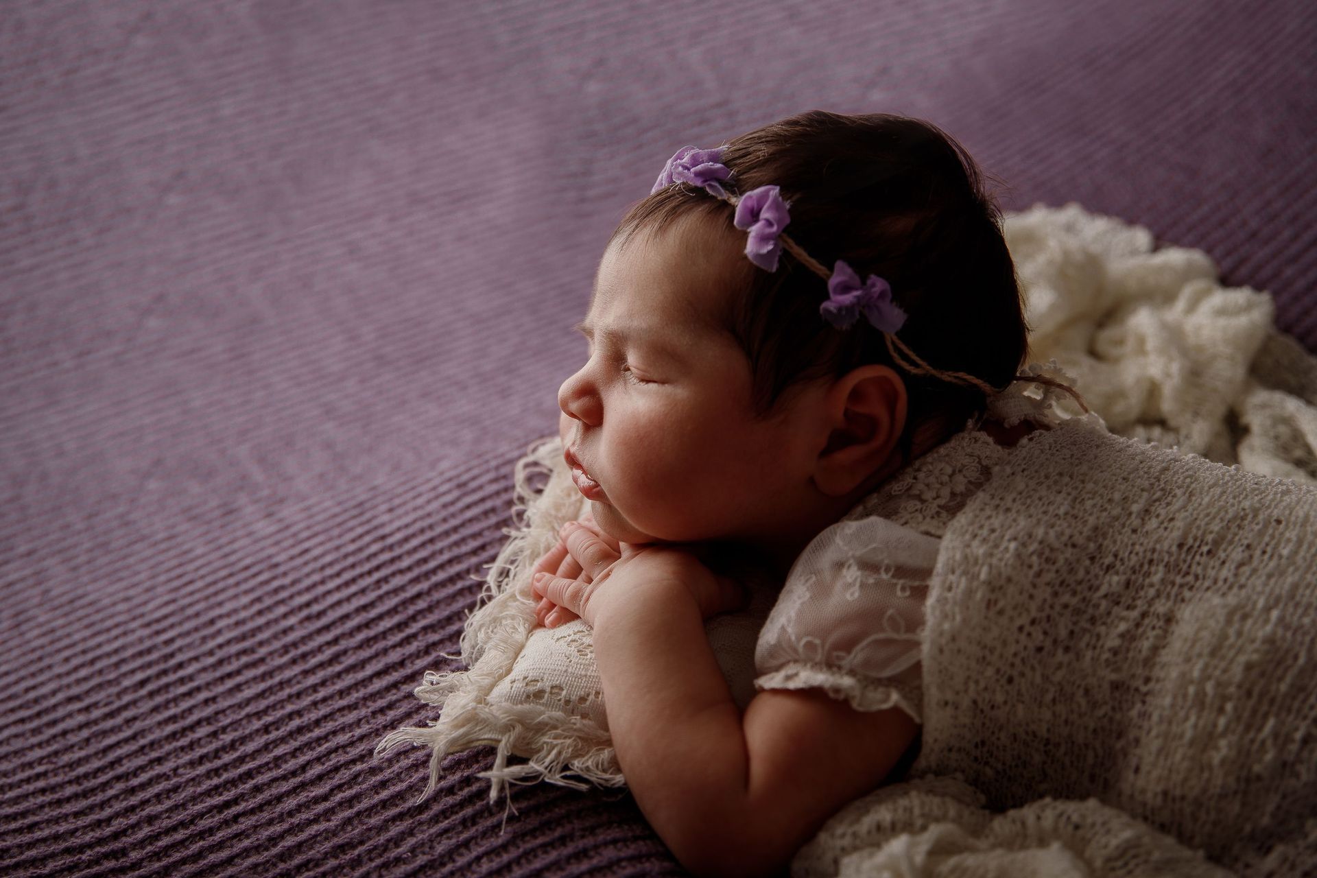 Bebé durmiendo sobre una manta morada, con los brazos cruzados y una diadema floral.