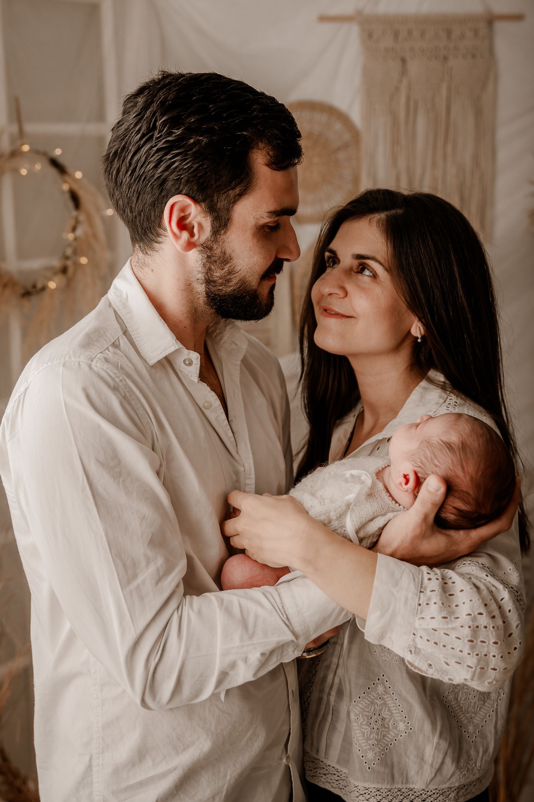 Una pareja sostiene a un recién nacido en una casa con iluminación tenue, mirándose con amor.