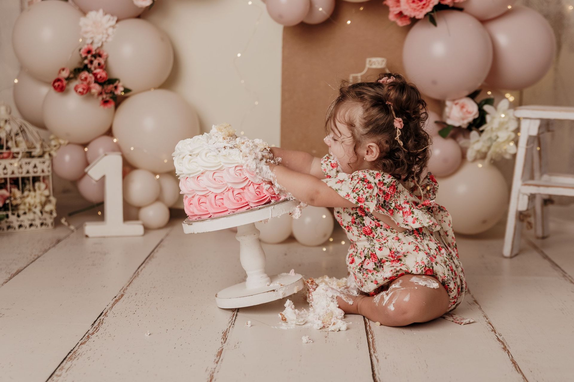Una niña pequeña con un atuendo floral destroza un pastel rosa glaseado en una decoración de cumpleaños con globos beige y un gran letrero con el número 