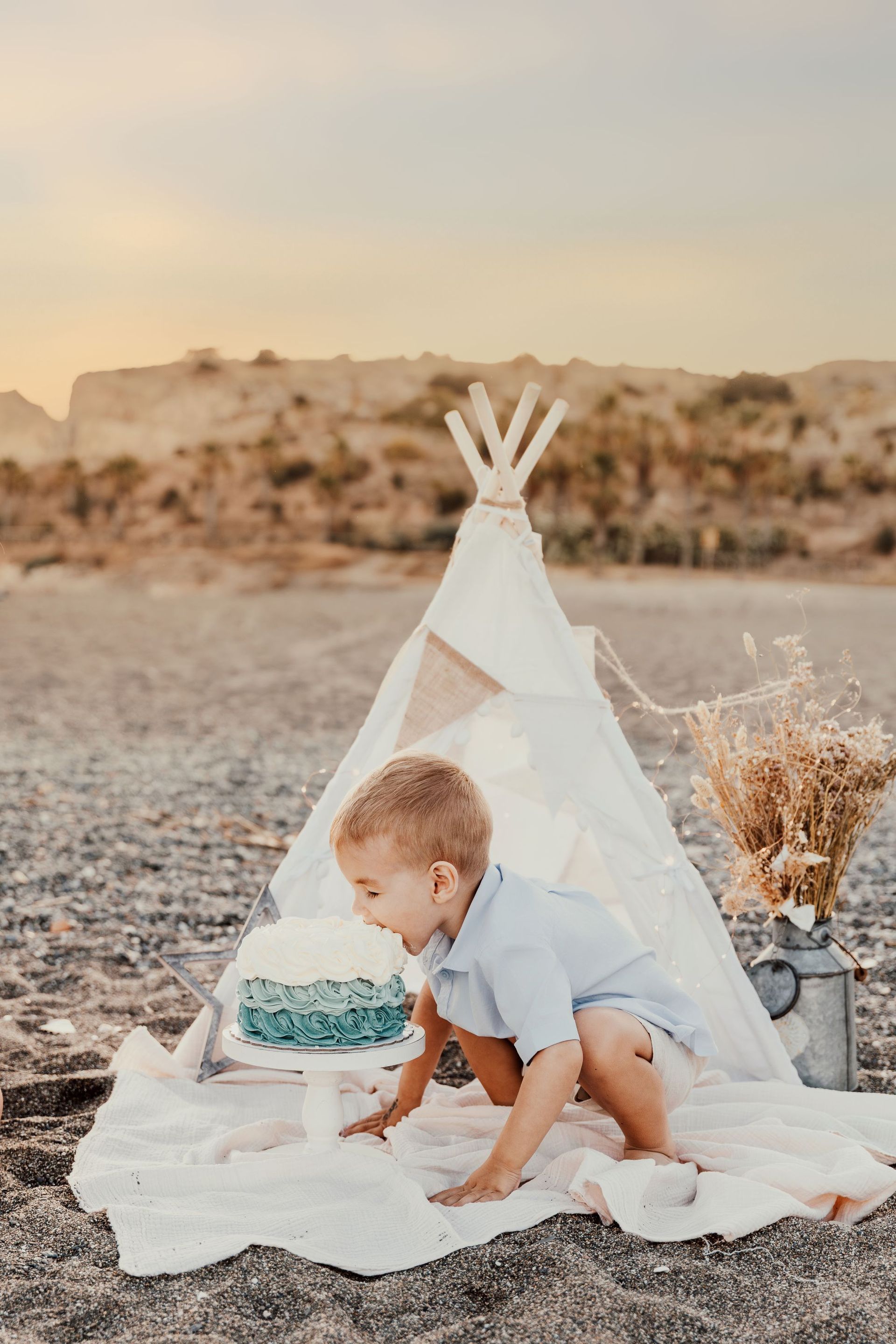 Niño arrodillado junto a una tienda tipi con un pastel sobre una manta en un entorno desértico.
