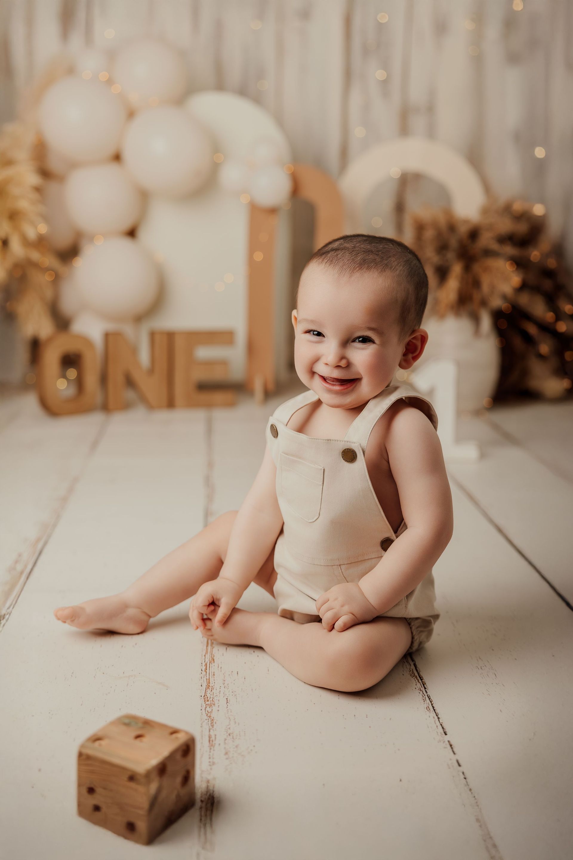Un bebé sonriente con un mono beige está sentado en un suelo claro con un bloque de madera, en un estudio decorado con globos.