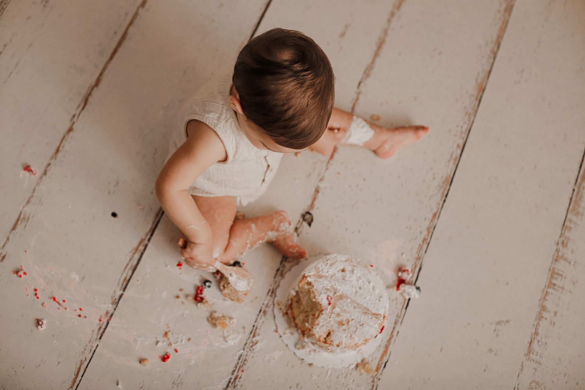 Un niño vestido de blanco juega con un pastel derramado en un suelo de madera blanca.