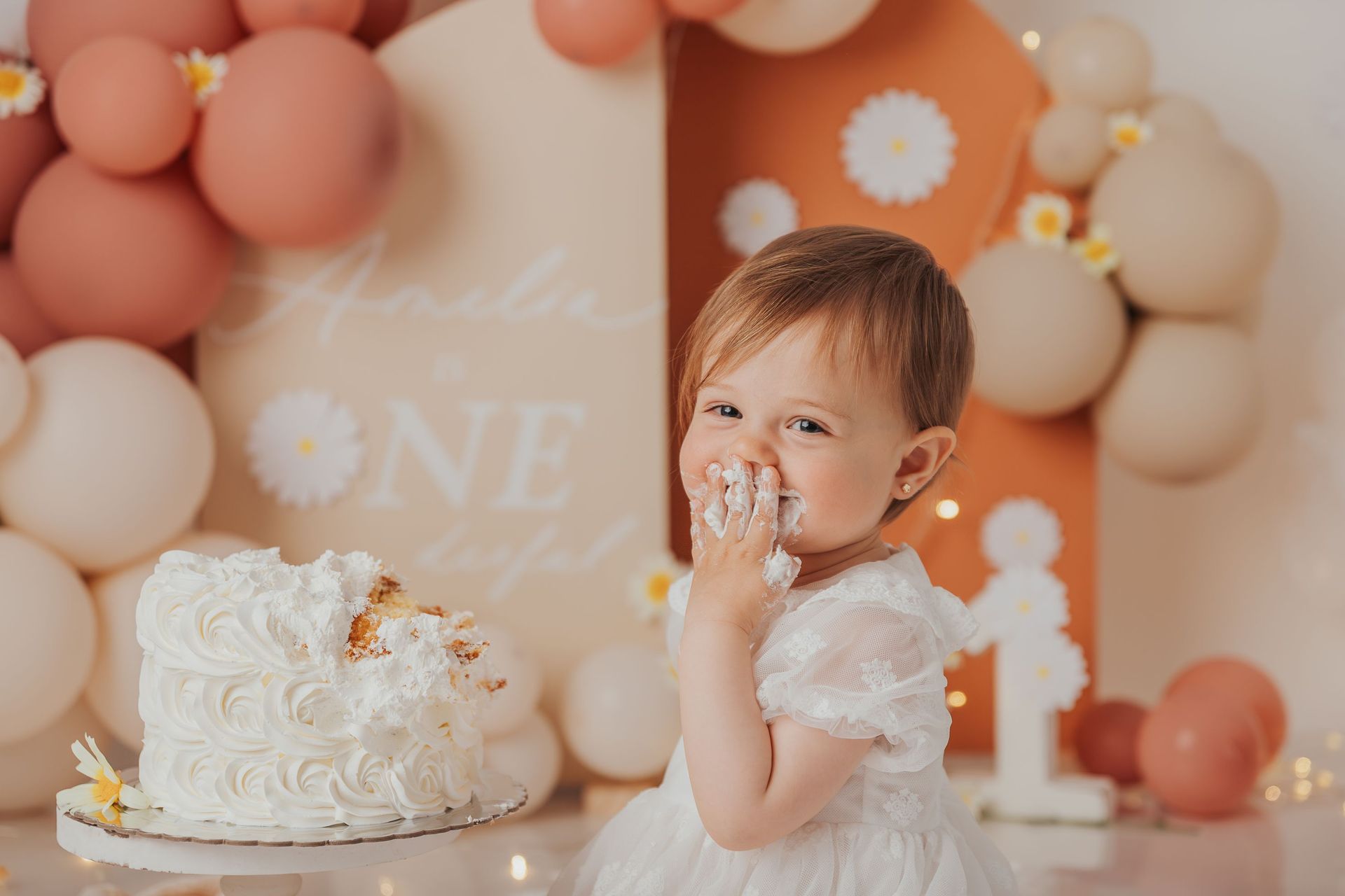 Una niña pequeña con un vestido blanco sonríe junto a un pastel de cumpleaños blanco y globos