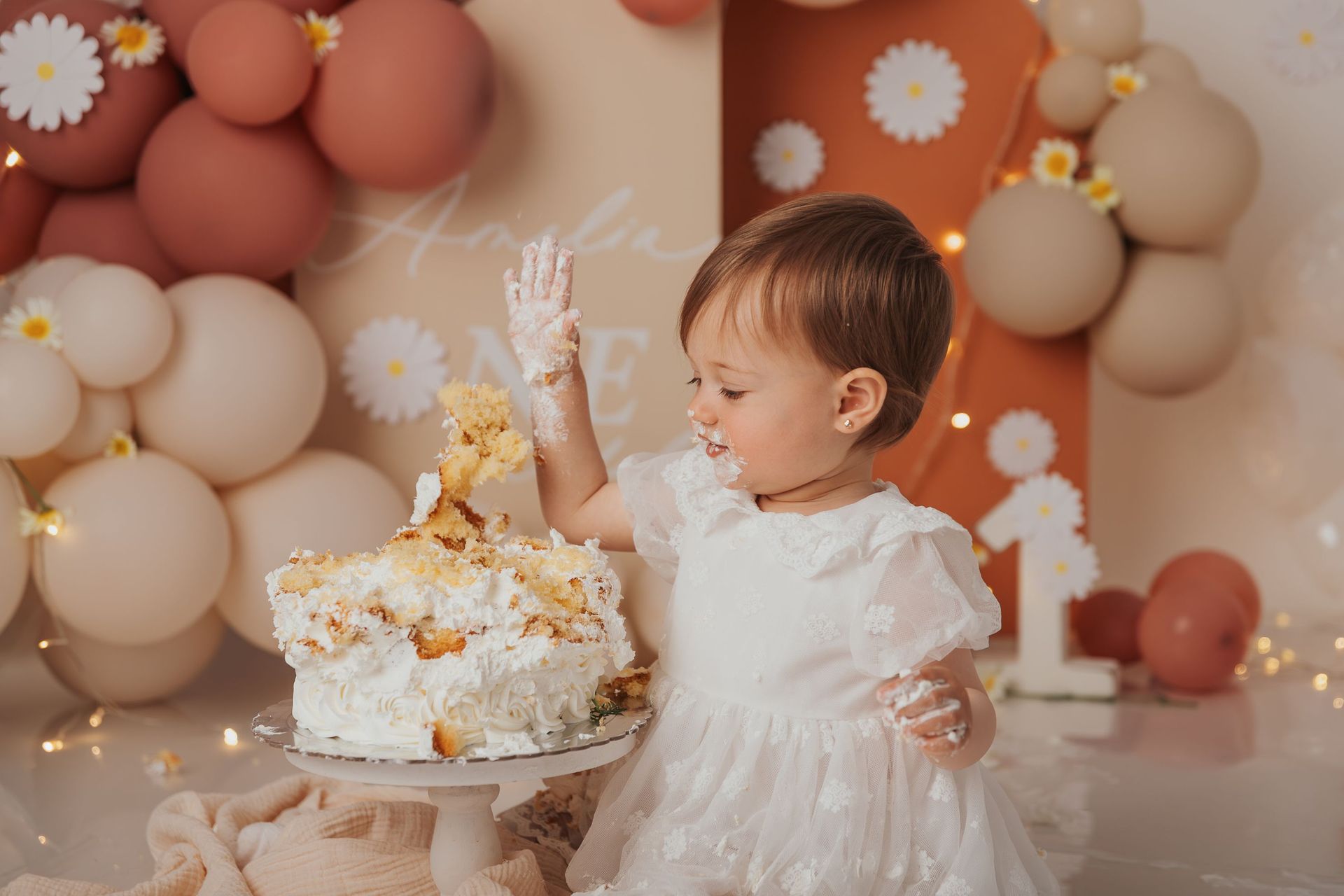 Una niña pequeña con un vestido blanco intenta alcanzar una tarta de cumpleaños con globos y margaritas de fondo.