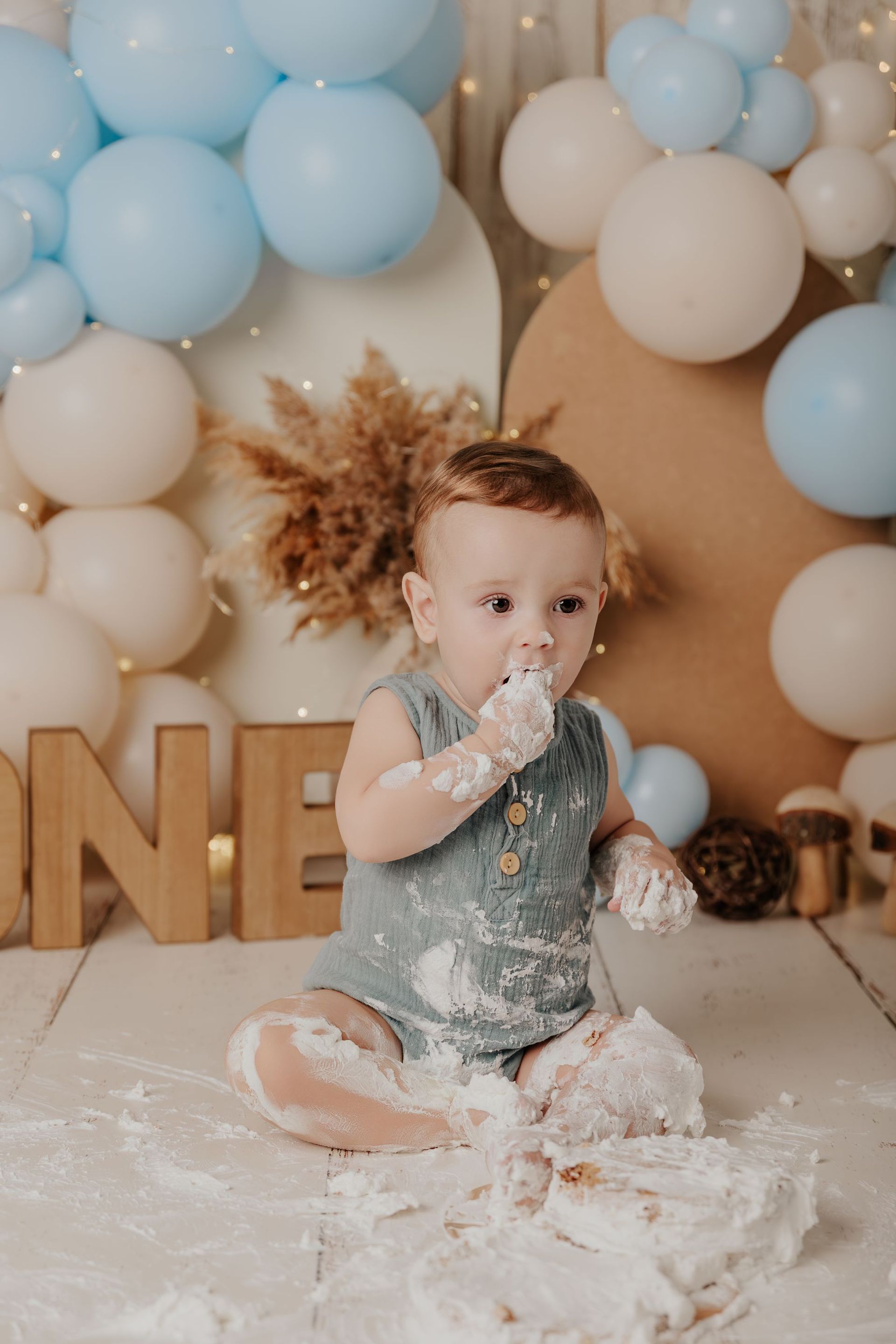 Un niño pequeño sentado en un escenario de pastel lleno de globos, cubierto de glaseado blanco y sosteniendo un dulce plateado.