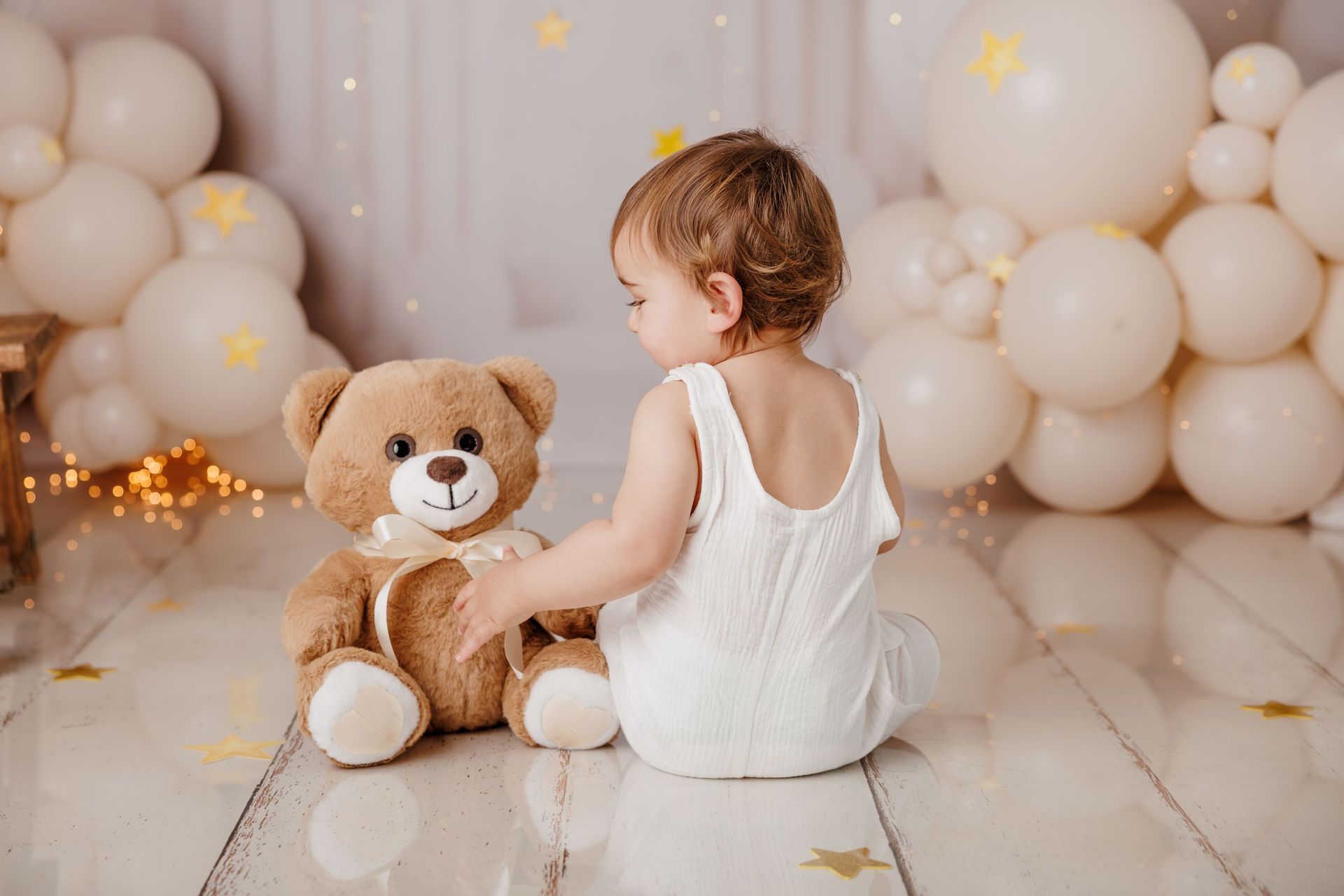 Una niña pequeña con un pelele blanco está sentada junto a un osito de peluche en una habitación llena de globos de colores pastel.