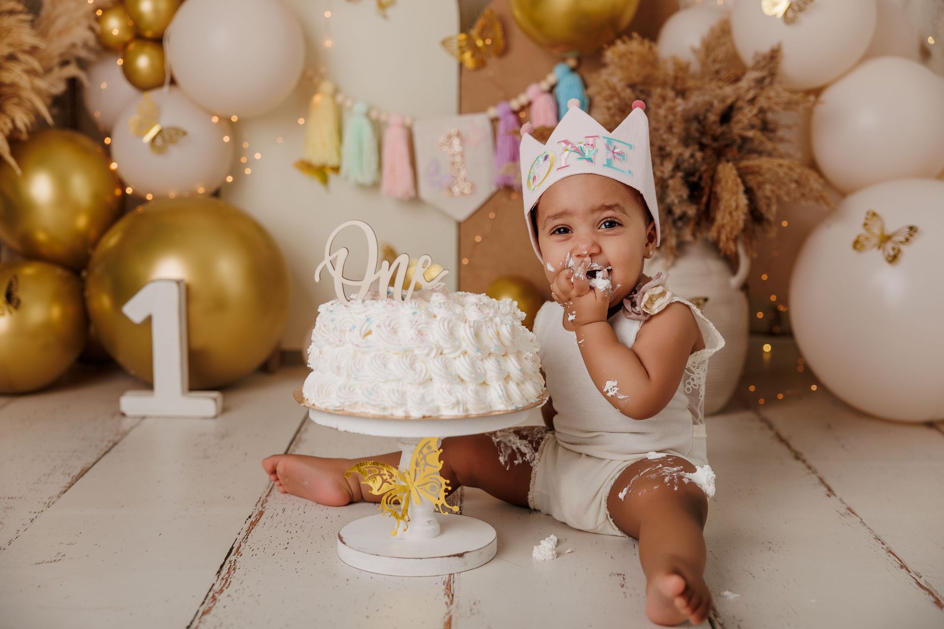 Bebé en su fiesta de primer cumpleaños con pastel, globos dorados y blancos y una corona, sonriendo y comiendo pastel.