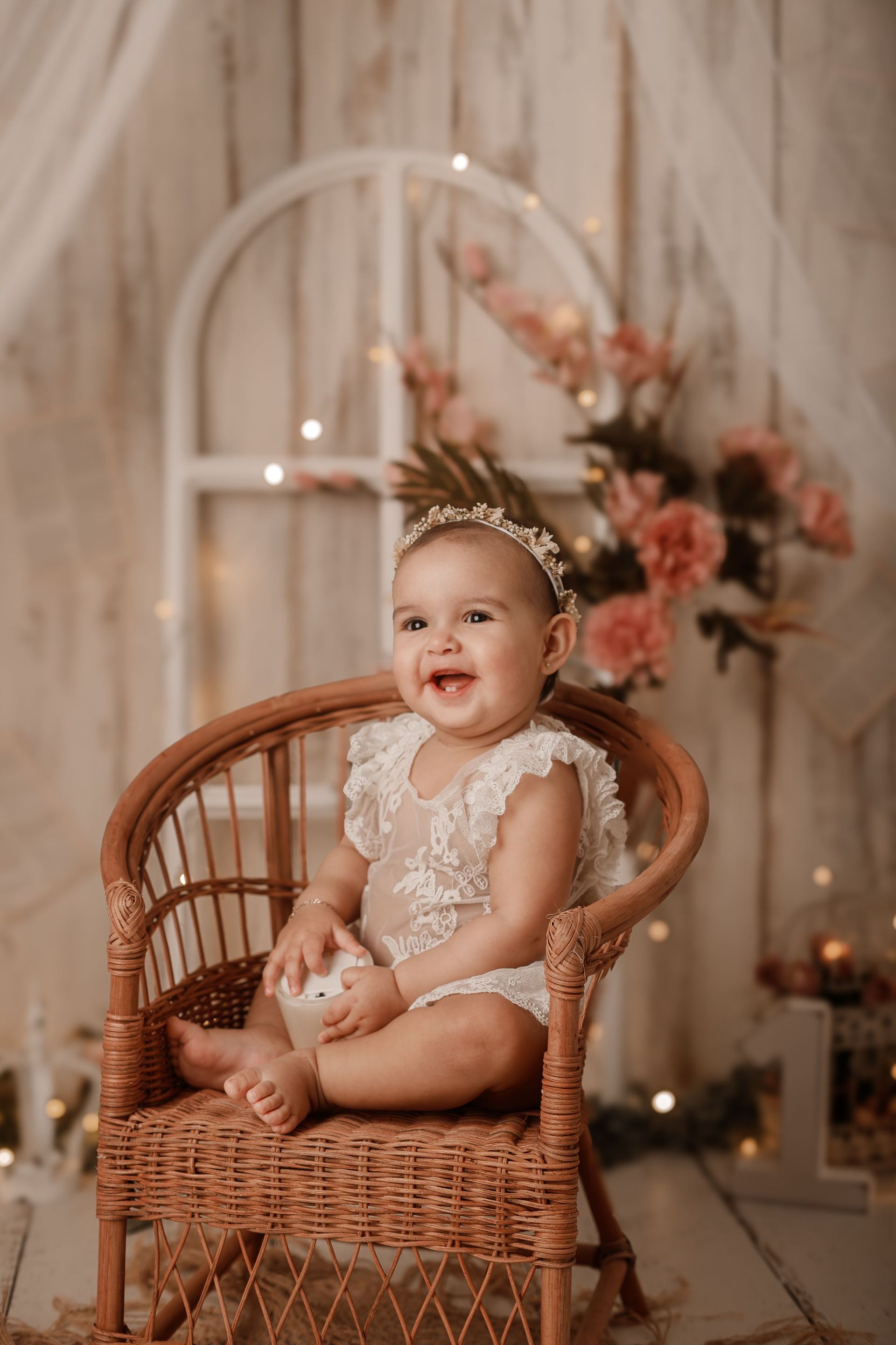 Bebé sonriente en una silla de mimbre, con un conjunto de encaje y una diadema floral, con un suave fondo floral.