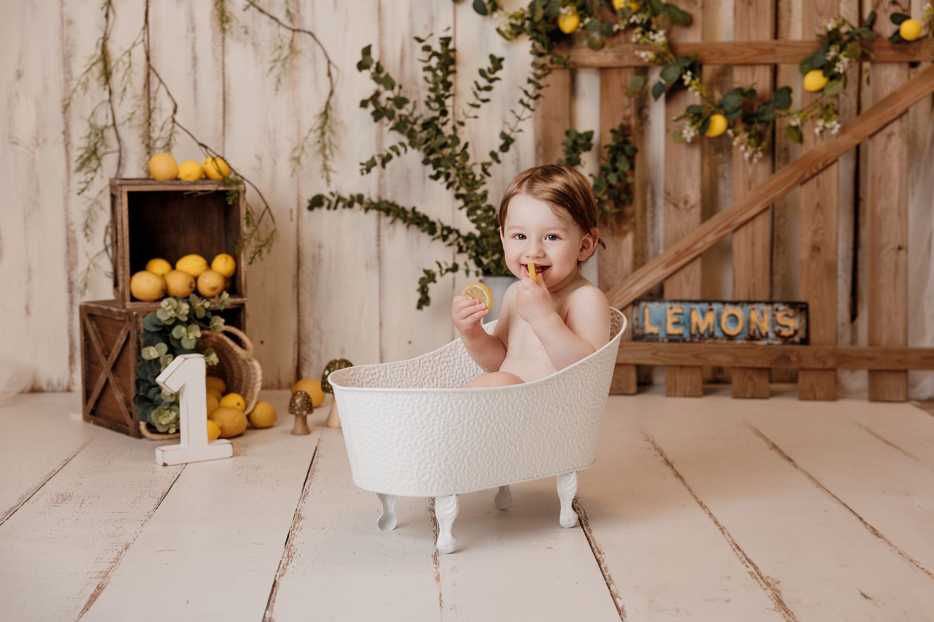 Un niño pequeño sentado en una bañera blanca sosteniendo un limón en un estudio rústico con temática de limones.