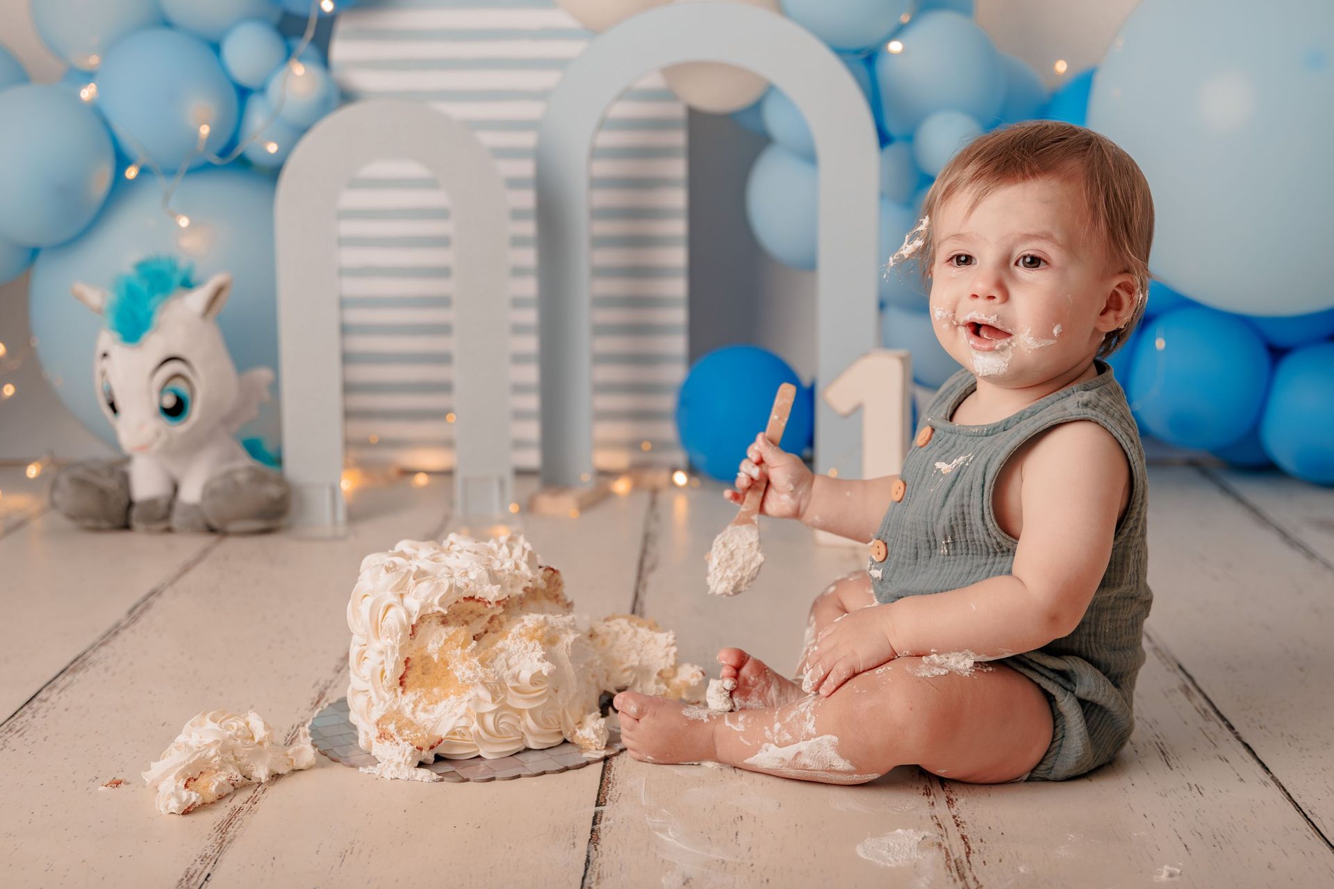 Un niño pequeño sentado en una sesión de fotos con pastel, globos azules y arcos blancos, sosteniendo un número uno.