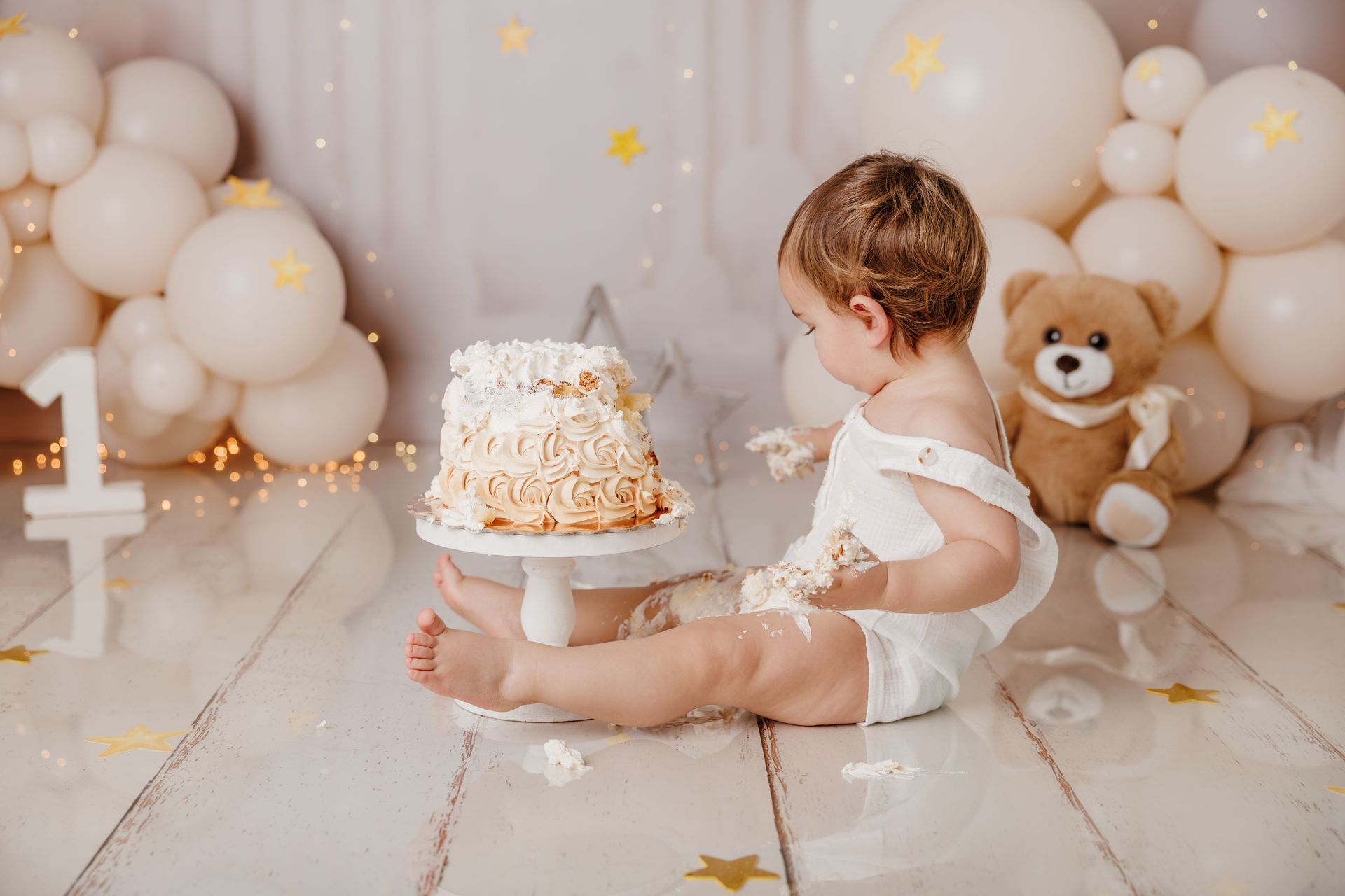Una niña pequeña vestida de blanco está sentada junto a una tarta en una fiesta de cumpleaños decorada con globos de colores pastel y un osito de peluche.