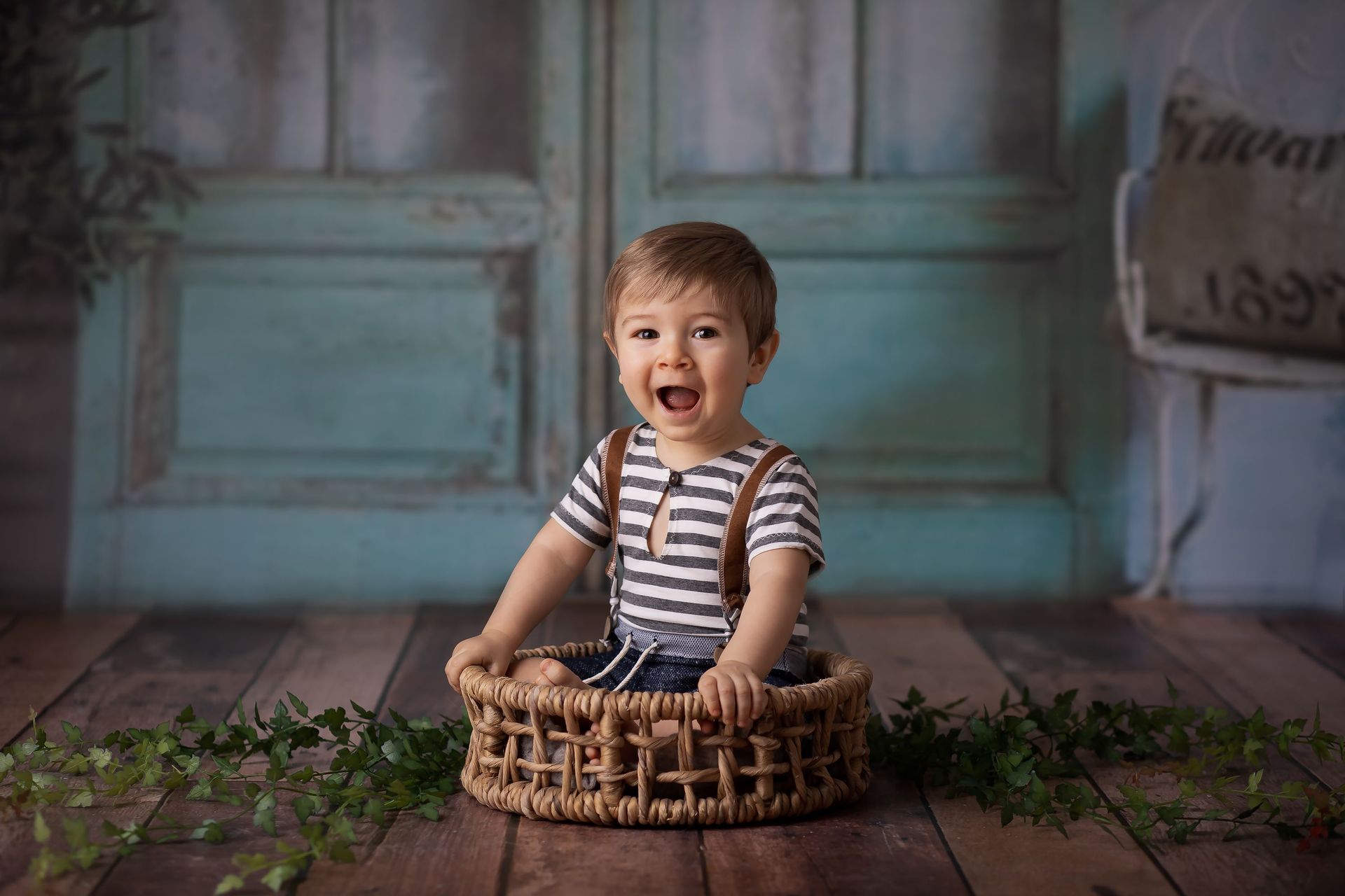 Niño sonriente sentado en una cesta de mimbre sobre un suelo de madera, con una camisa de rayas en una habitación rústica.