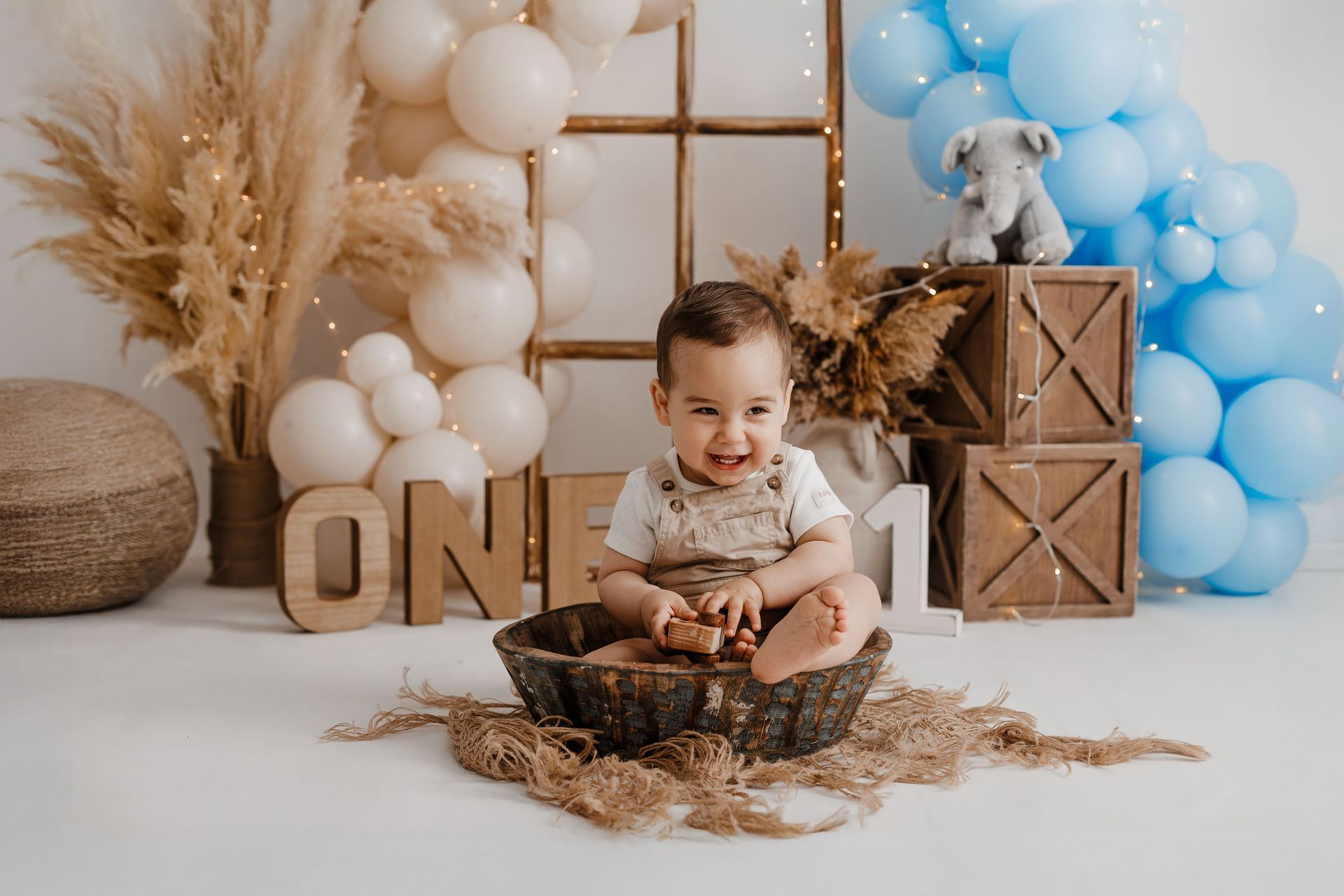 Un bebé sonriente sentado en una cesta con globos blancos y azules y bloques de madera en un entorno festivo.