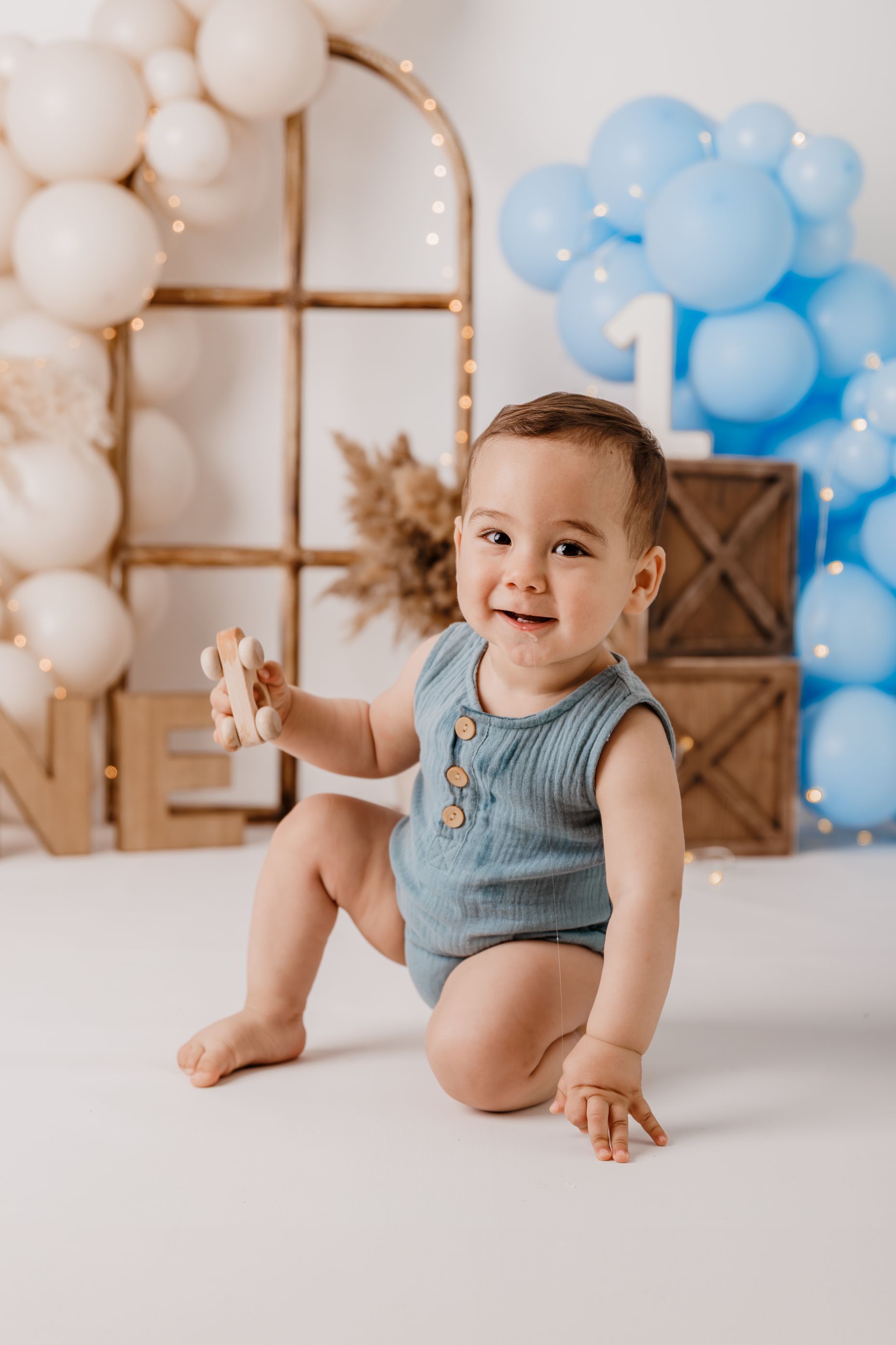 Bebé sonriente con un pelele azul sentado en un suelo blanco con decoraciones de globos al fondo.