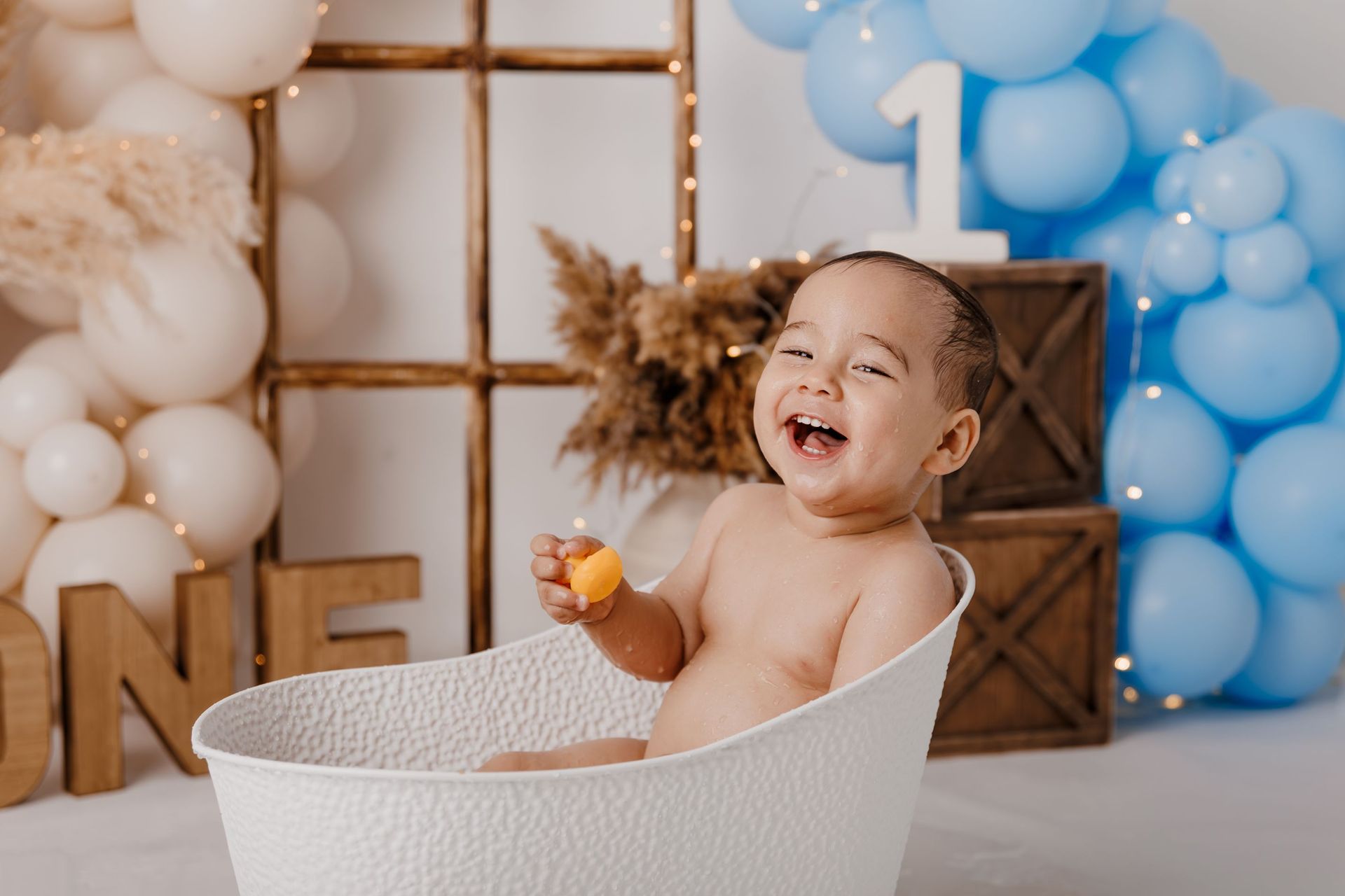 Un bebé riendo en una cesta blanca en una fiesta de primer cumpleaños azul y blanca con globos y decoraciones.