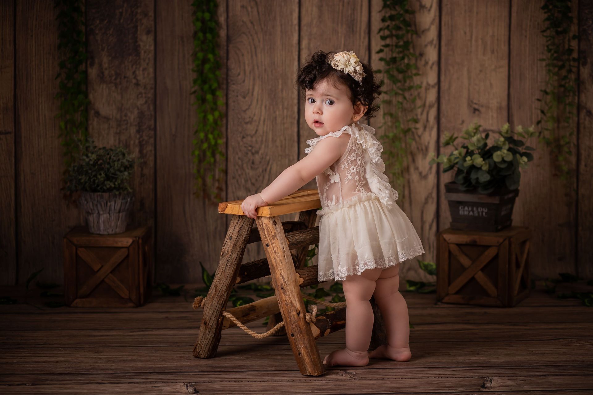 Una niña pequeña con un vestido color crema está de pie junto a un taburete de madera en un estudio rústico