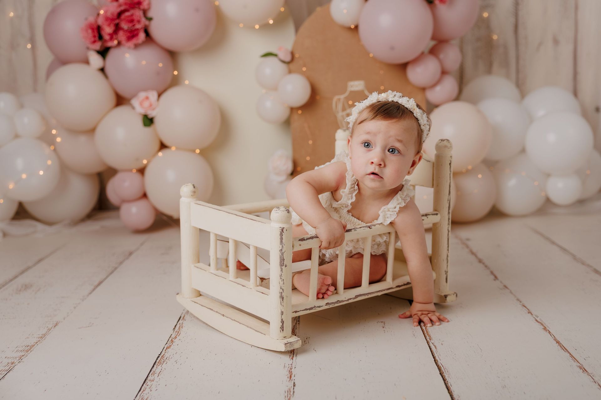 Un bebé sentado en una pequeña cuna blanca entre globos de colores pastel en un ambiente de estudio acogedor.