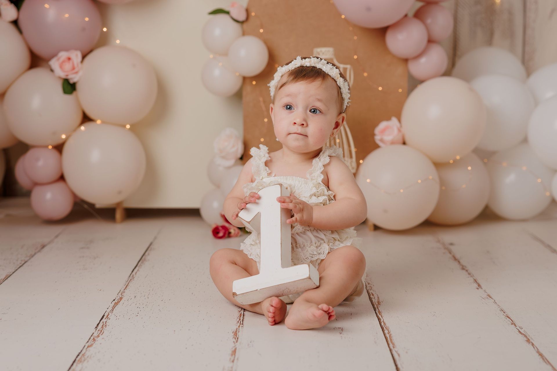 Un niño pequeño sentado entre globos rosas y blancos, sosteniendo un adorno para pastel con el número uno sobre un suelo de color pastel.