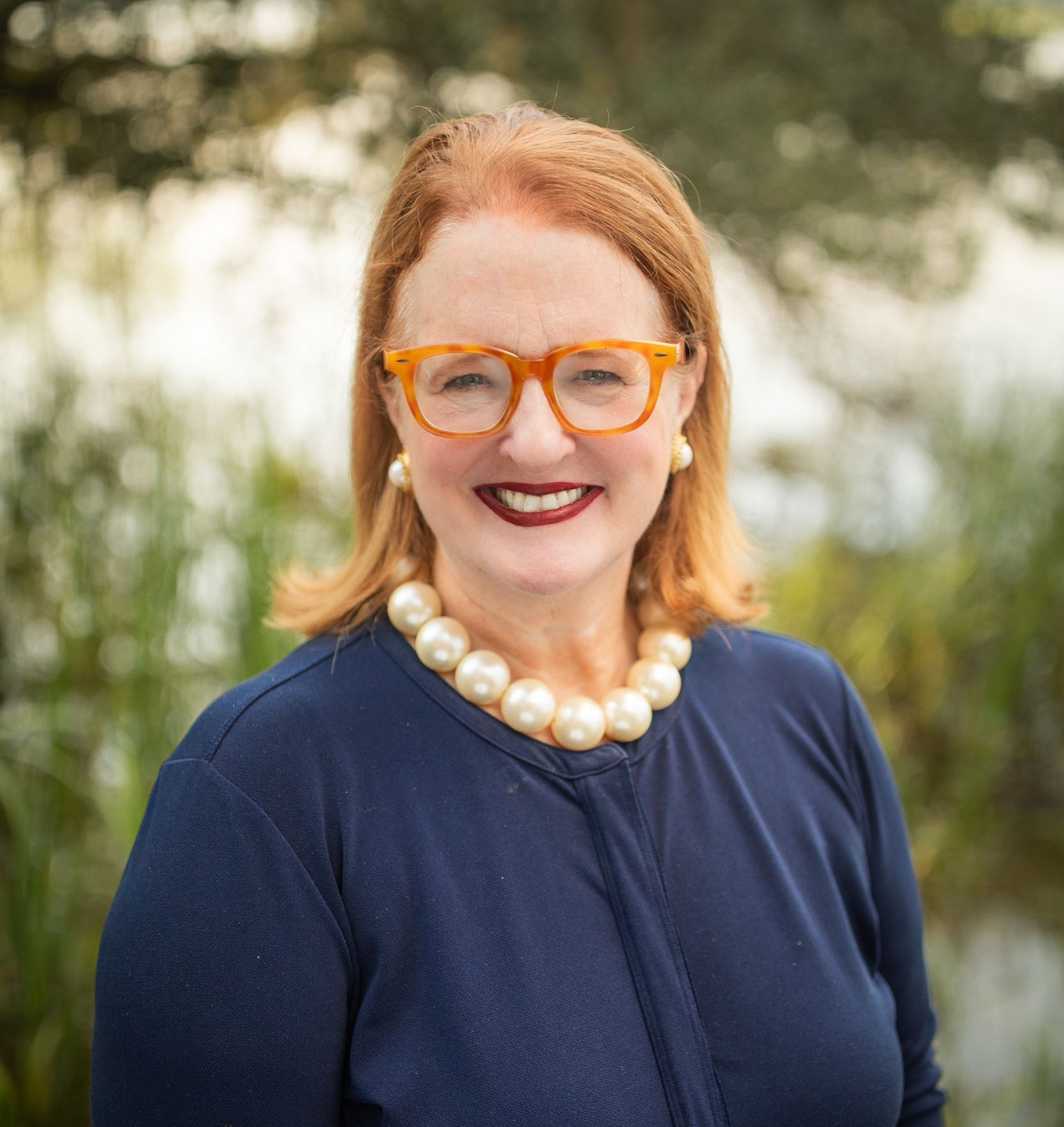 A woman wearing glasses and a black jacket is standing in the woods.
