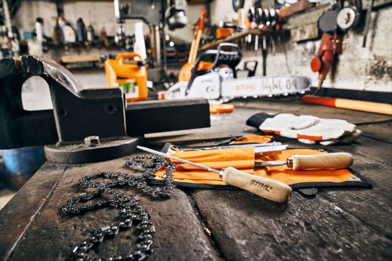 Eine rustikale Werkbank mit einer Kettensäge, einer Metallkette, Schärfwerkzeugen in einer orangefarbenen Rolle und Arbeitshandschuhen.