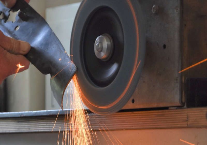 Eine Person schärft ein Rasenmähermesser aus Metall an einer rotierenden Schleifscheibe, wodurch ein Schauer hellorangener Funken entsteht.