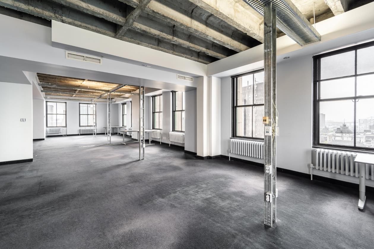 Espace de bureaux vide, de style industriel, avec de grandes fenêtres, des poutres apparentes et de la moquette grise.