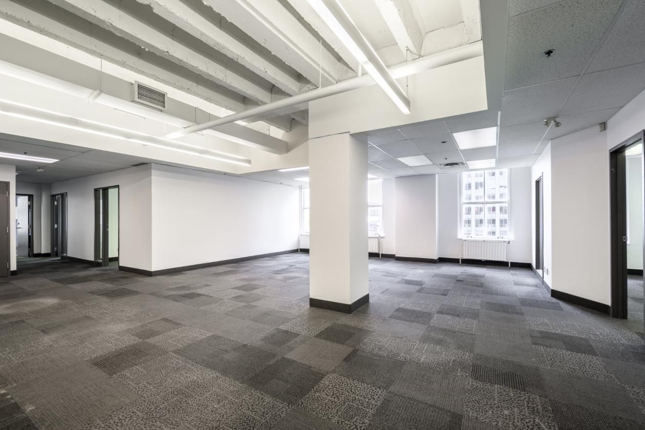 Espace de bureaux vide et décloisonné avec moquette grise à motifs, murs blancs et grandes fenêtres.