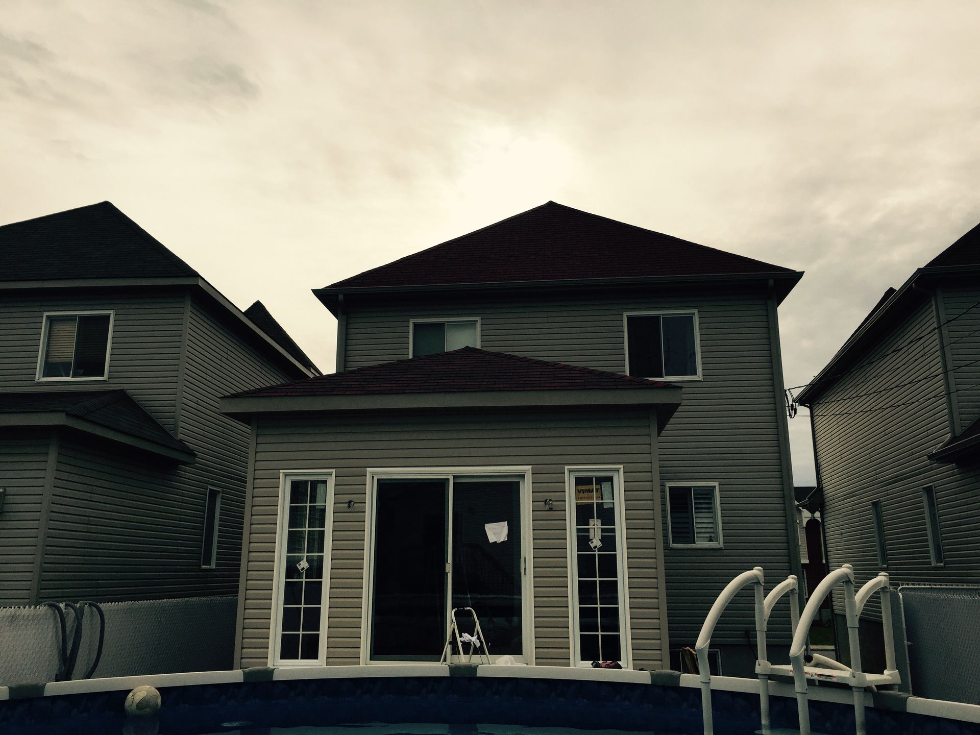 Vue arrière d'une maison avec piscine, bardage gris, toit marron, ciel nuageux.