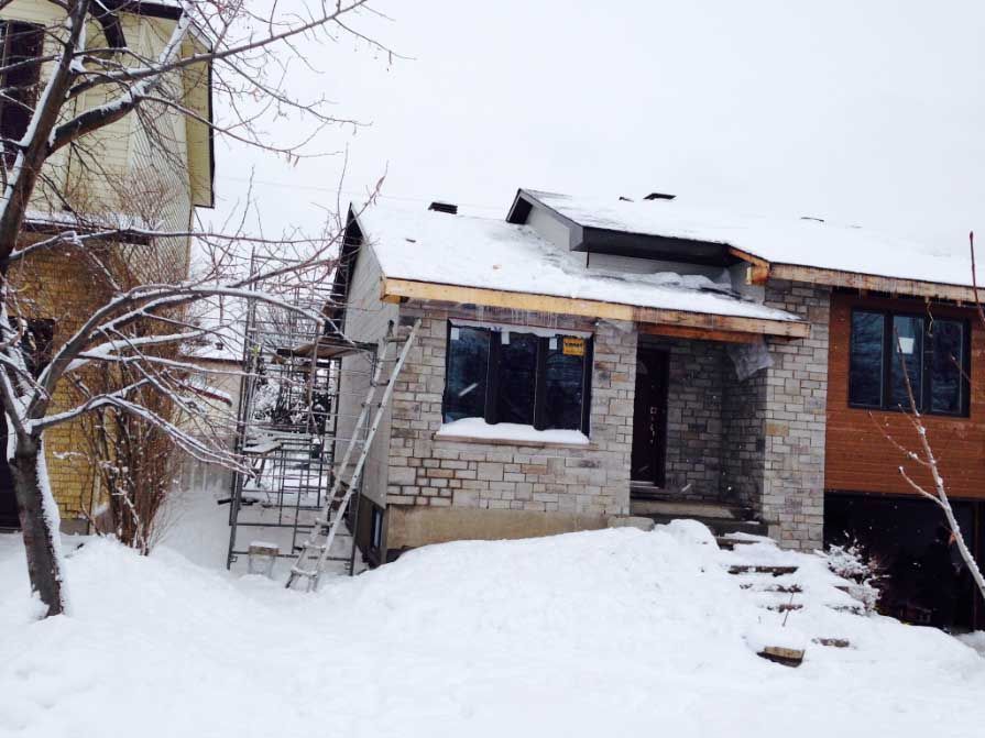 Maison en construction en hiver ; façade en pierre, marches enneigées, échafaudages, toiture inachevée.