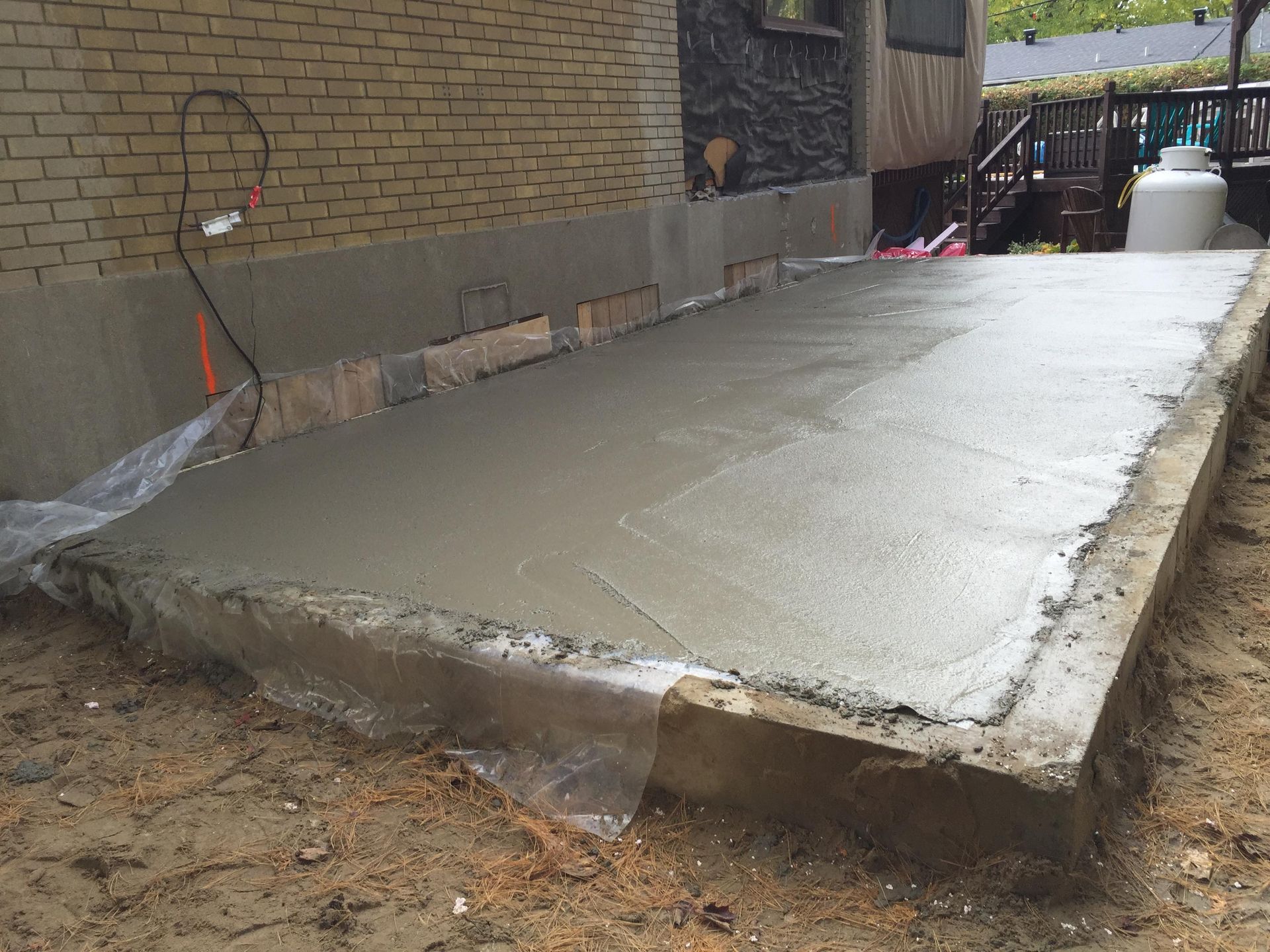 Terrasse en béton fraîchement coulée contre un bâtiment en briques ; bâche en plastique visible.