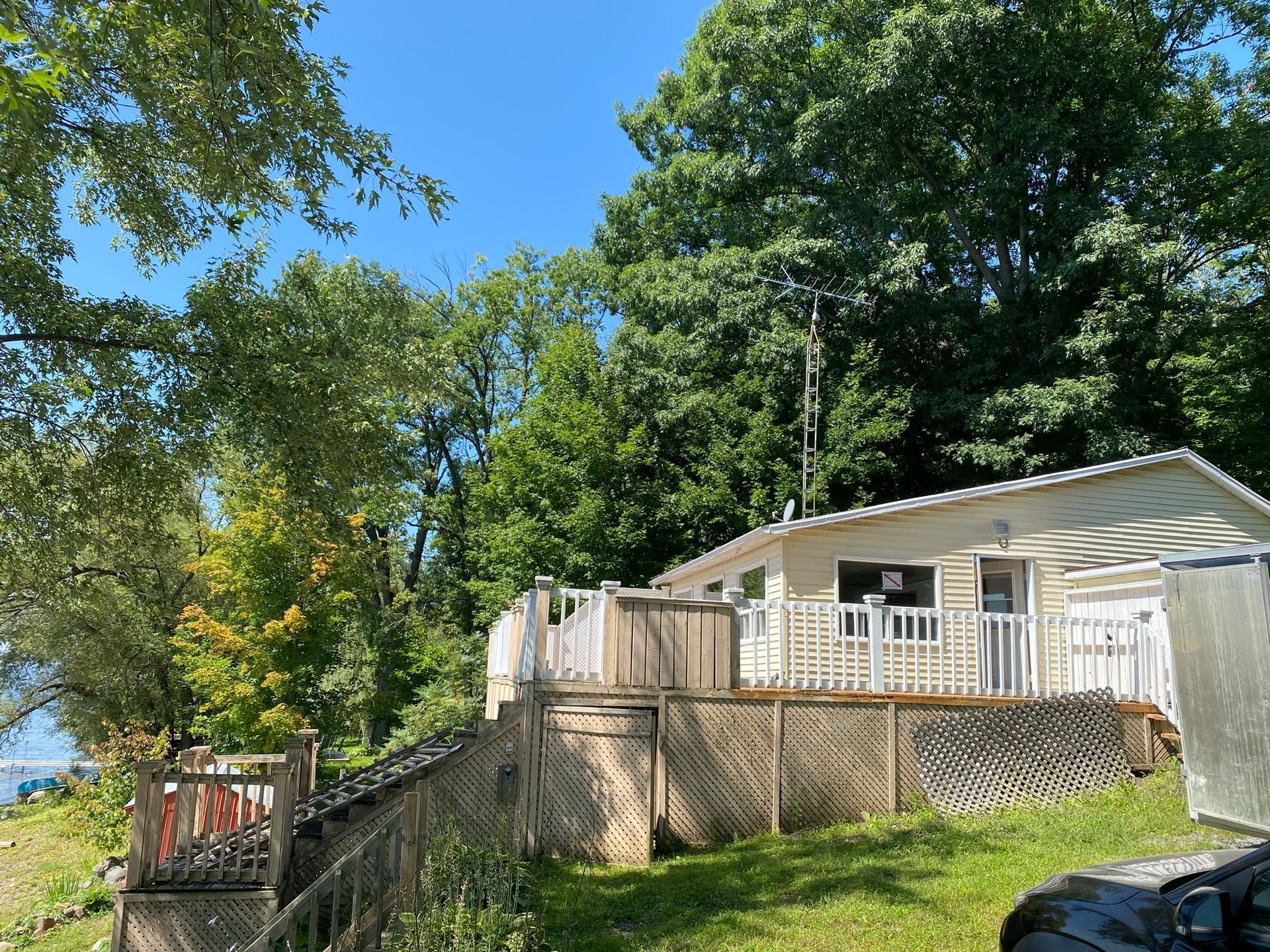 Chalet avec terrasse blanche, entouré d'arbres verts sous un ciel bleu, près de l'eau.